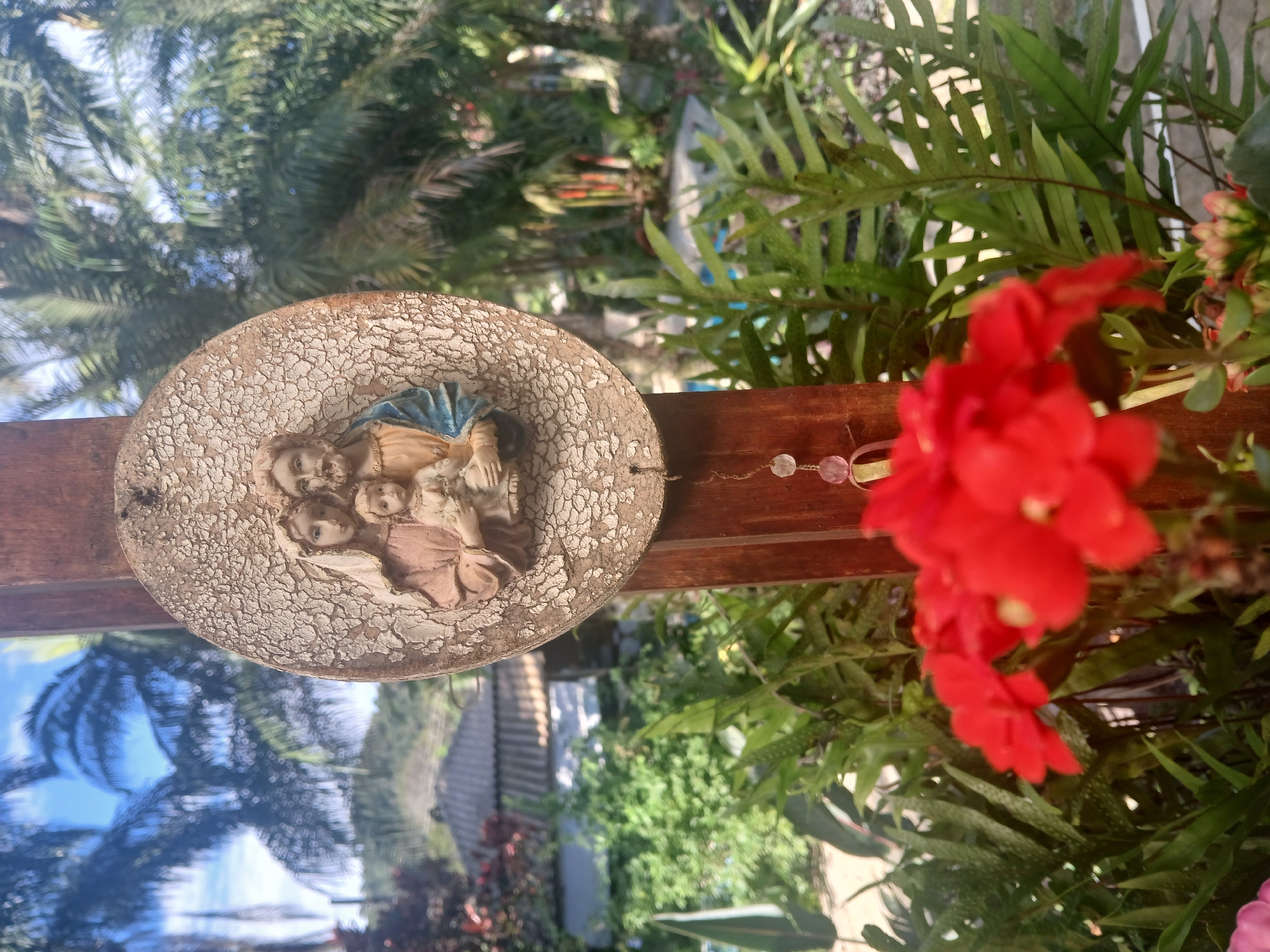 The image is a close-up, eye-level shot of an oval plaque depicting the Holy Family: Mary, Joseph, and baby Jesus. The plaque is mounted on a wooden post in an outdoor setting. The plaque has a cracked, distressed finish with raised figures painted in muted colors. Mary wears a pink robe and a blue veil, holding the baby. Joseph wears a blue garment and has a beard.
Below the plaque, a beaded string hangs, with pink and clear beads and a small, yellow object. In the foreground, out of focus, are vibrant red flowers. Behind the plaque and flowers, there is lush green foliage, including ferns. Further in the background, palm trees with fronds are visible against a bright blue sky with some clouds. To the left, the roof of a building with gray tiles can be seen, surrounded by more greenery.
The scene suggests a garden or a veranda area, possibly in a tropical or subtropical climate due to the palm trees. The overall atmosphere is serene and religious. The specific location in the city of Rio de Janeiro, Brazil, cannot be determined from the visual information alone.