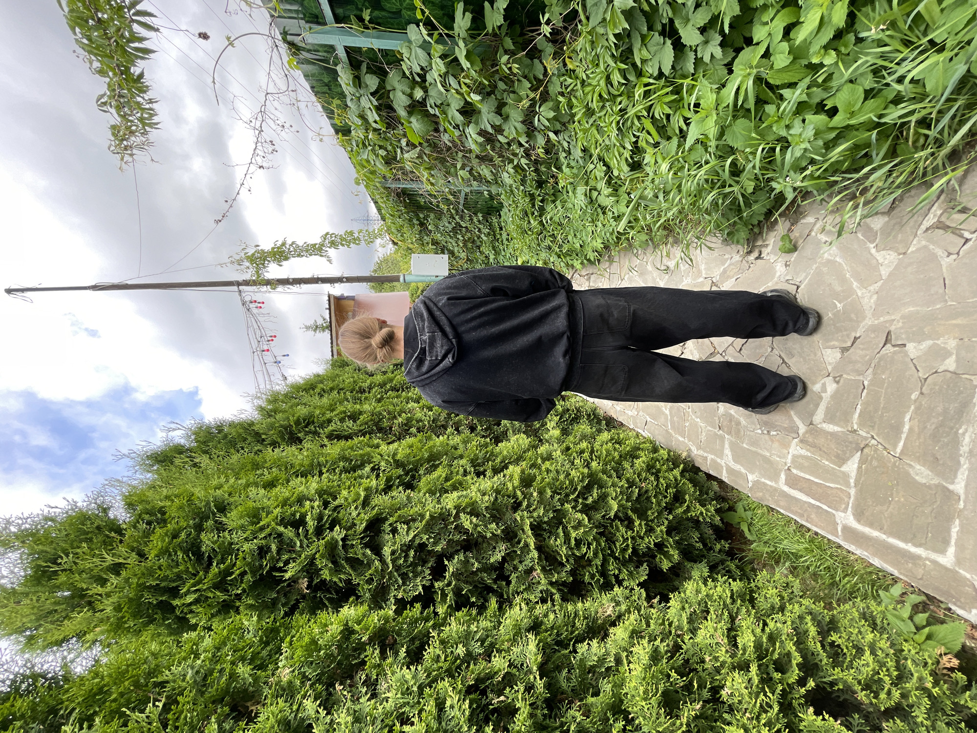 A person wearing black clothes walks on a stone path between a tall, green hedge and a fence covered in vegetation. The location is Kommunarka, Russia. Overhead, the sky is cloudy with glimpses of blue. A utility pole with red and blue decorations is visible in the background.