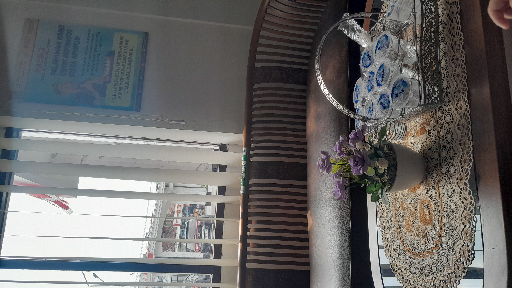 The image shows a seating area with a brown couch and a coffee table. On the coffee table, there is a white pot with purple artificial flowers and a silver basket filled with small cups of bottled water. A decorative lace doily is placed under the flower pot. In the background, through a window with vertical blinds, an outdoor scene is visible, including a building with a red sign that appears to say "SRC". There is also a black SUV parked outside. A poster is hanging on the wall to the right of the window, with text in Indonesian that includes the phrase "PELAYANAN KAMI TIDAK DIPUNGUT BIAYA APAPUN" which translates to "OUR SERVICES ARE FREE OF CHARGE." A red and white flag is also visible through the window. The overall impression is of a waiting area in a public or government building, possibly in the city of Tasikmalaya, Indonesia, given the text on the poster.