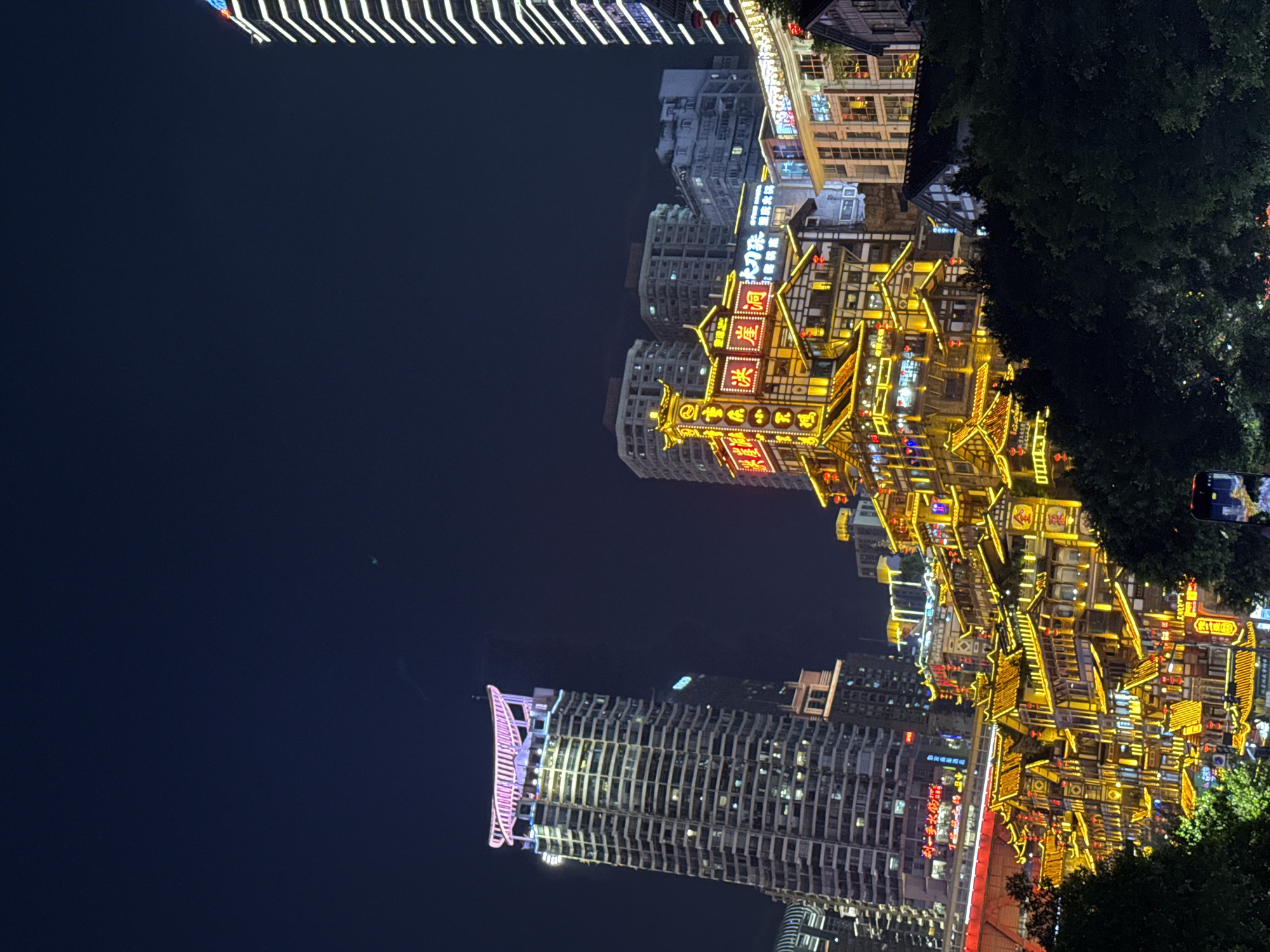 This media file captures a vibrant night cityscape in Yuzhong District, Chongqing, China, showcasing a striking blend of traditional and modern architecture. The scene is dominated by a multi-tiered complex of traditional Chinese stilt-house buildings, brightly illuminated with warm yellow, orange, and red lights. These structures feature intricate roofs, balconies, and decorative elements, suggesting a historical or cultural district. Prominent neon signs with Chinese characters are visible on these buildings, including "洪崖洞" (Hongyadong) on a large red sign, clearly identifying the famous landmark. Another sign also displays "重慶" (Chóngqìng).
In stark contrast, several modern high-rise buildings rise in the background and to the left, their windows and facades glowing with various patterns of artificial light. One tall skyscraper on the left stands out with distinctive vertical white illumination and a curved top section adorned with pinkish-purple lights. The dark, night sky provides a dramatic backdrop, making the extensive artificial lighting stand out. Dense, dark foliage from trees can be seen along the right edge of the frame, adding a touch of nature to the urban environment.
The time of day is clearly night, evidenced by the dark sky and the reliance on artificial illumination. While no specific individuals are clearly visible, the lively lighting and nature of the buildings suggest a bustling urban area, likely a popular tourist destination or commercial hub. The overall atmosphere conveyed is one of energy, cultural richness, and urban grandeur, highlighted by the juxtaposition of ancient and contemporary styles.