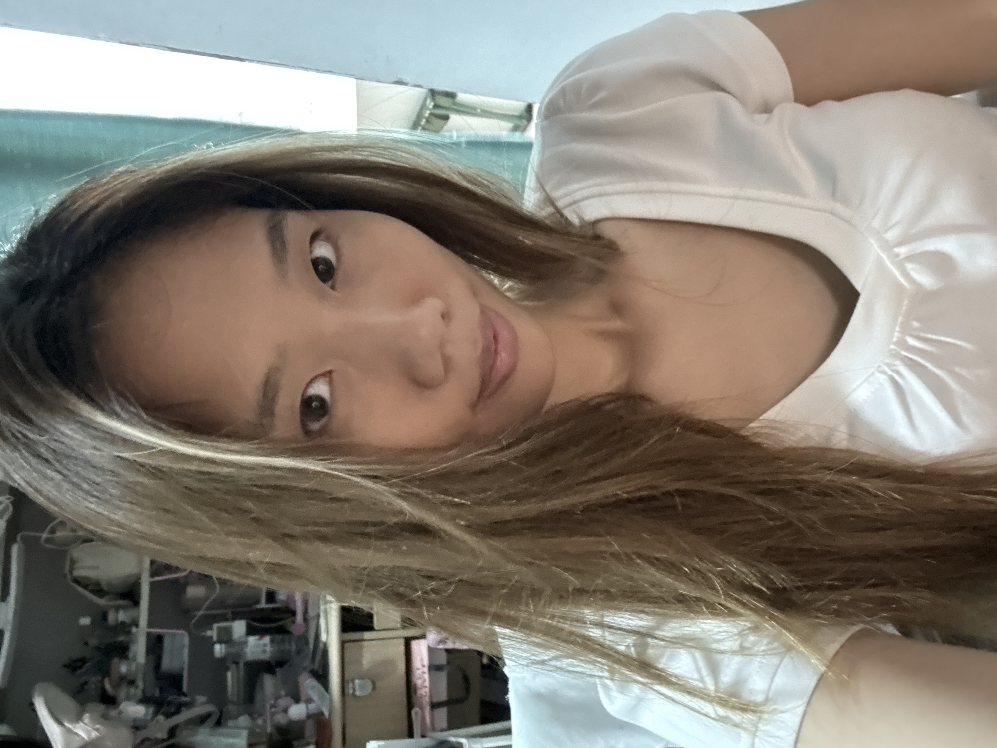 A close-up, medium shot shows a young woman with light brown, shoulder-length hair that is slightly messy, framing her face. She is wearing a white V-neck t-shirt. Her right eye is visible, looking directly at the camera, while her left eye is partially obscured by her hair. Her lips are slightly parted. The background is blurred, but shelves filled with various items, including bottles and boxes, are visible behind her to the left. To the right, a teal curtain and a white wall are present, suggesting an indoor setting, possibly a room in Nan'an District, China.