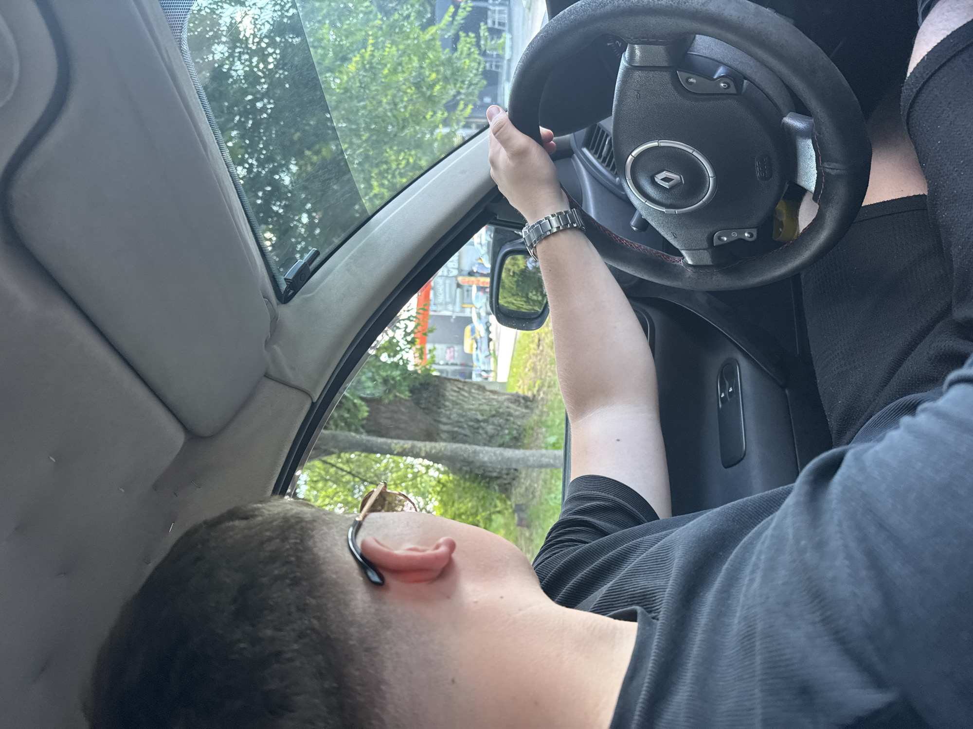 The image shows a person driving a car in Chisinau, Moldova. The driver, viewed from the side and from behind, is wearing a black t-shirt and sunglasses. Their left hand is on the steering wheel, which has a Renault logo in the center. The car interior is visible, including the dashboard and the passenger side window. Outside the car, trees and other vehicles are visible, suggesting an urban environment. The lighting indicates it is daytime.