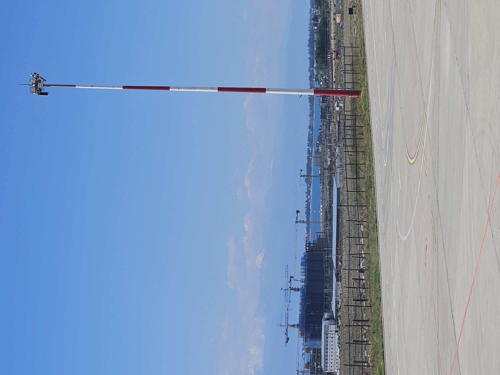 The image depicts an outdoor scene in Choktal, Kyrgyzstan, on a clear day with a bright blue sky. In the foreground, a paved surface, likely an airport tarmac, is visible with white and red markings. To the right, a tall, red and white striped pole with lights and equipment at the top stands prominently. In the middle ground, a fenced area with barbed wire encloses a grassy strip. Beyond the fence, a body of water is visible, with a construction site featuring multiple cranes and unfinished buildings on the left. In the background, a mountain range with snow-capped peaks forms a distant backdrop. The overall environment appears to be an airport or industrial area near a body of water and mountainous terrain.