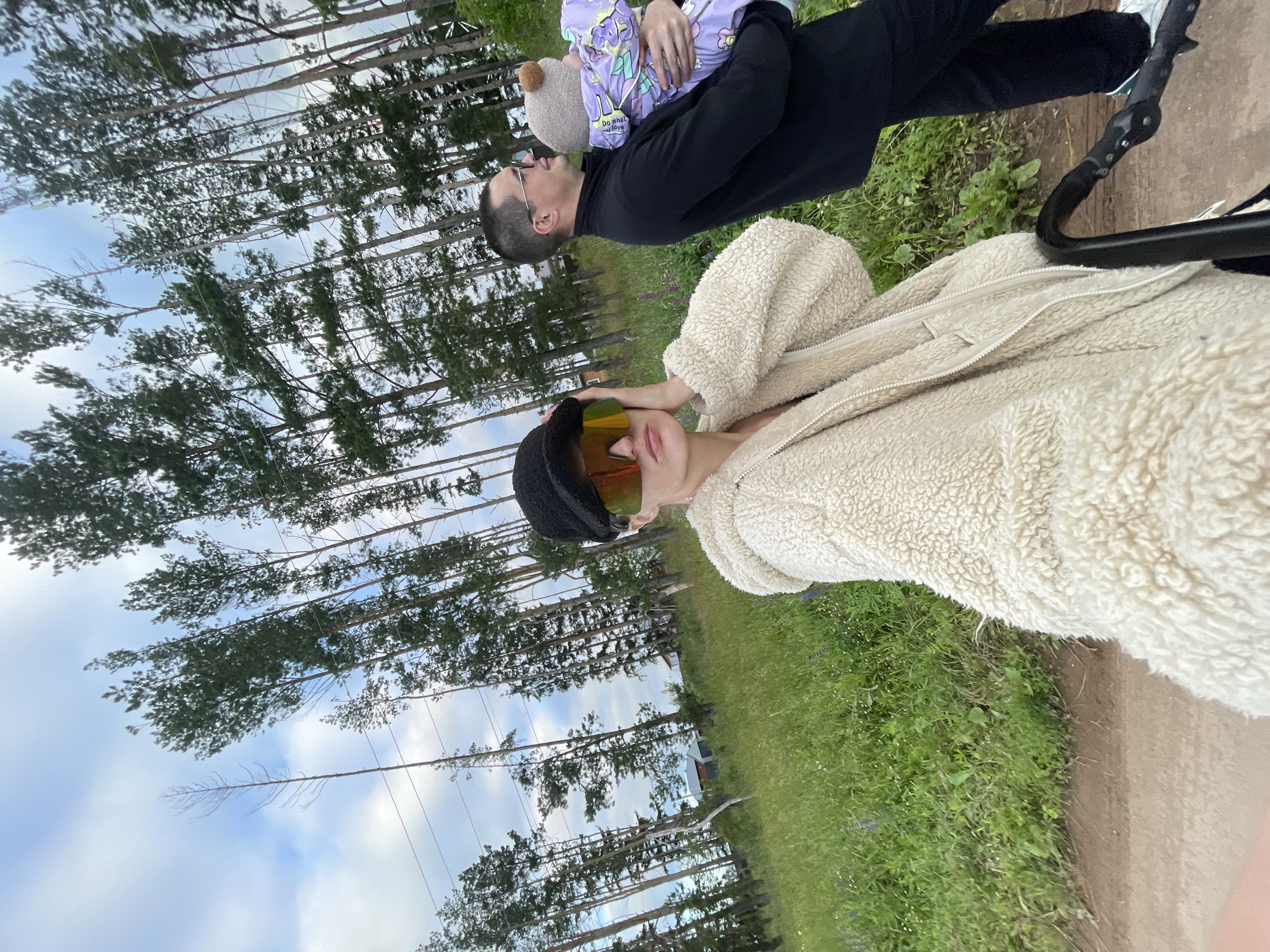 The image shows two people and a baby outdoors in a grassy area with trees in the background. The scene appears to be a portrait taken from a low angle, looking up towards the subjects.
In the foreground, a person wearing a fluffy cream-colored jacket and sunglasses is looking towards the camera. They are wearing a black hat. Behind them, another person is holding a baby. The baby is wearing a purple outfit with cartoon characters and is holding a small teddy bear. The person holding the baby is wearing glasses and a black long-sleeved shirt. The background consists of a line of tall, thin trees against a cloudy sky. The ground is covered in green grass with some small white flowers. The overall lighting suggests it is daytime.
There is some text visible on the baby's outfit which reads "do what you love". The location context is Podshivalovo, Russia. The image has a casual and affectionate feel, with the focus on the family unit and the natural outdoor setting.