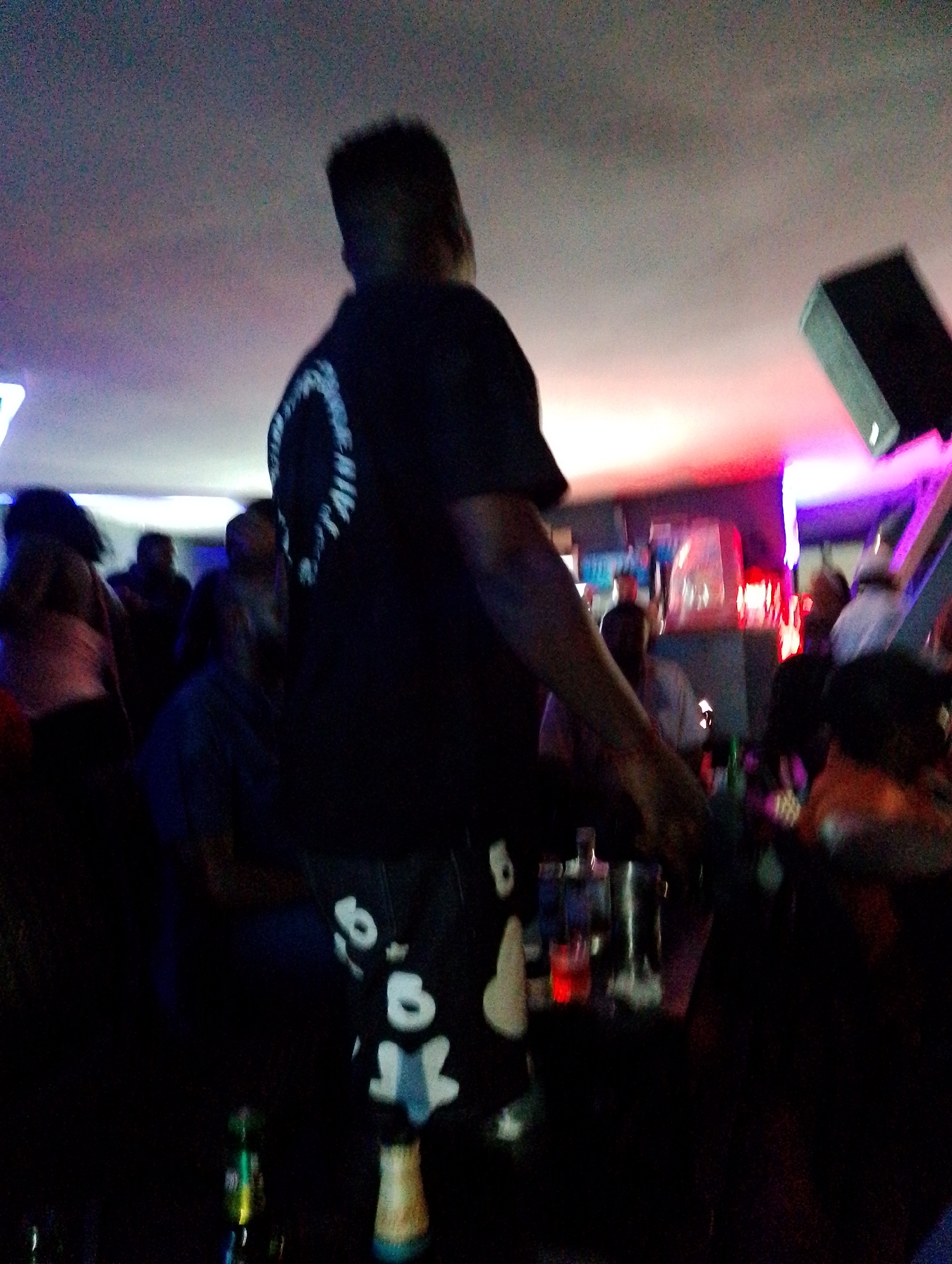 The image depicts the interior of a crowded nightclub or bar in Kubwa, Nigeria. The subject in the foreground is a person wearing a dark-colored shirt with a graphic design on the back, and pants with stylized white shapes. The person appears to be standing near a bar area. In the background, there are other individuals, some of whom appear to be socializing or dancing. Various bottles and bar equipment are visible on the bar counter. The lighting in the venue is dim, with artificial colored lights illuminating the scene. A black speaker is mounted on the wall.