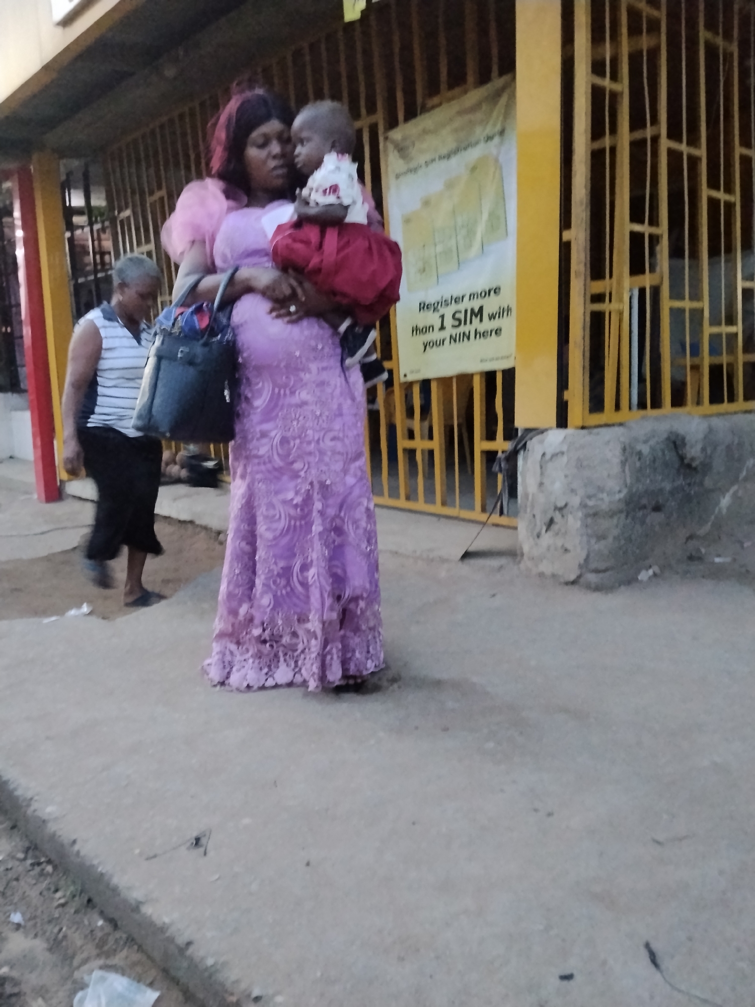 A woman in a pink dress is holding a baby in her arms. She is standing on a sidewalk in front of a building with yellow bars. A sign on the building reads "Register more than 1 SIM with your NIN here". Another person wearing a striped shirt is walking in the background. The location is identified as Ura Nta Umuarandu, Nigeria.