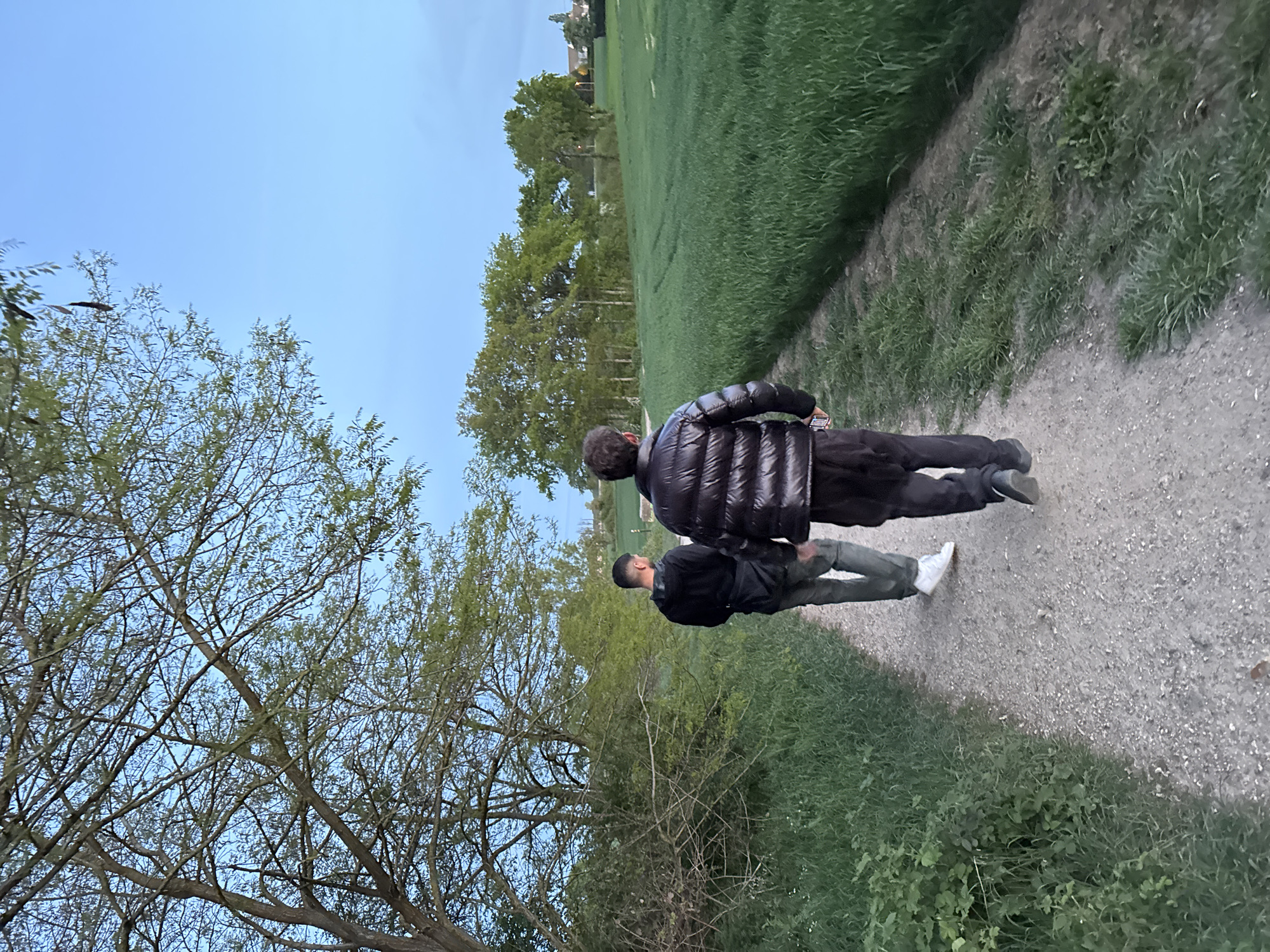 The image, taken in Gometz-le-Châtel, France, shows two men walking along a gravel or dirt path through a natural landscape. The photo is vertically oriented, with the path leading away from the viewer towards the upper right.
The subjects are two men, viewed from behind. The man in the foreground wears a dark, possibly black, puffy jacket and dark pants, with short, dark hair. The second man, positioned slightly behind and to the left of the first, is dressed in a dark jacket, light-colored (possibly olive green or grey) pants, and white athletic shoes.
The scene is dominated by a clear, bright blue sky at the top. To the left of the path, a dense arrangement of trees with green foliage suggests a wooded area or tree line. To the right of the path, a wide expanse of vibrant green grass stretches out, resembling a field or park. In the far background, faint structures, likely buildings or houses, are visible on the horizon, hinting at a nearby settlement.
The activity depicted is a leisurely walk. The bright, clear sky indicates pleasant weather, and the overall lighting suggests it is daytime, possibly late afternoon or early evening. No text is visible in the image. The setting portrays a serene, semi-rural environment.