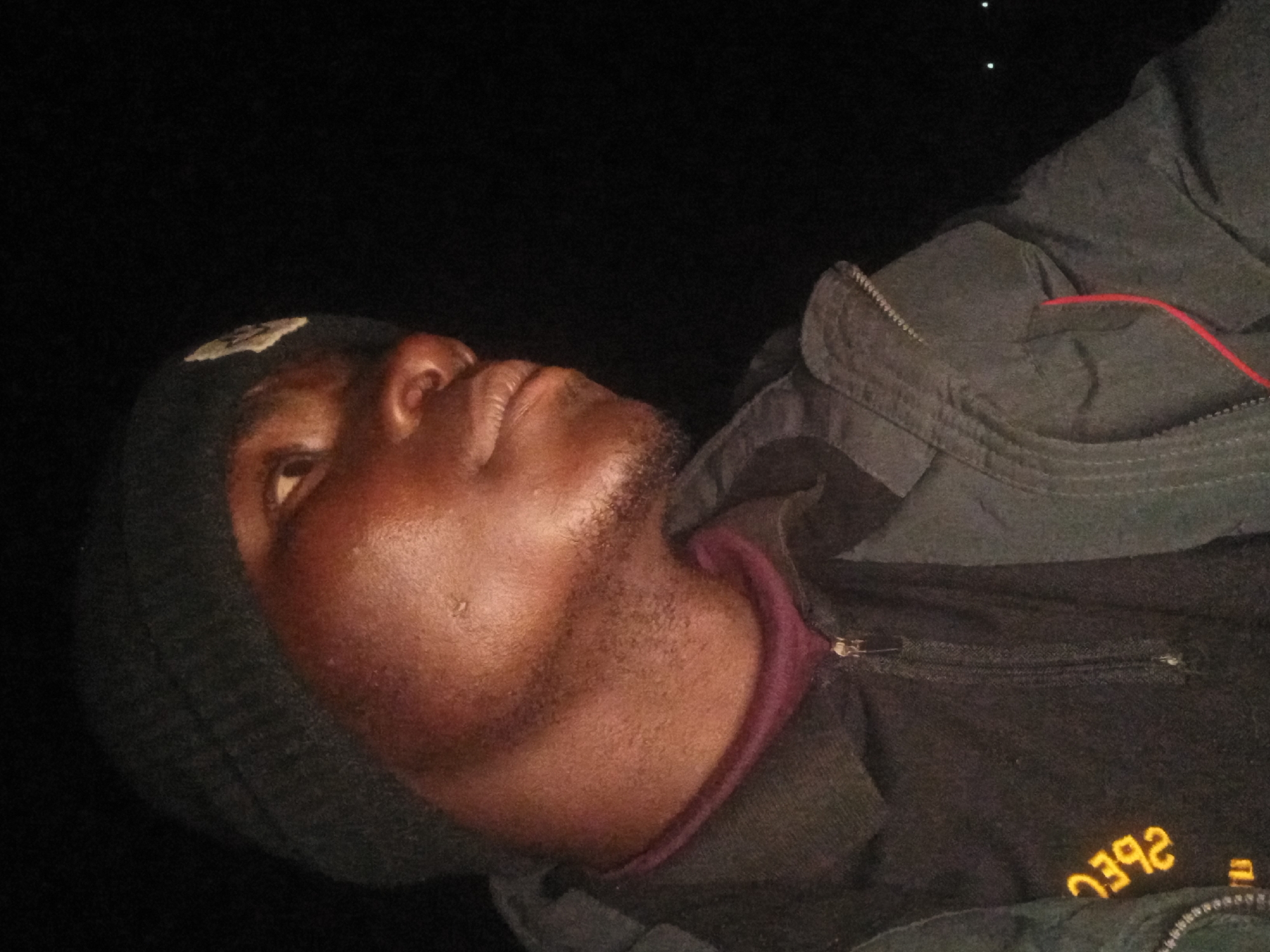 A close-up, profile shot of a young Black male wearing a dark beanie and a dark jacket over a black shirt with yellow lettering appears to be taken at night, possibly outdoors in Kwang, Nigeria. The lighting is artificial and casts shadows on his face, highlighting his cheekbone and the bridge of his nose. He has a slight stubble and is looking off to the right of the frame. The background is completely dark, with only a few faint lights visible in the distance, suggesting an outdoor, possibly rural or less urbanized setting. The jacket has a zipper and a red string detail. The yellow lettering on his shirt is reversed, appearing as "EBL2" from his perspective. The overall impression is a candid, casual photograph taken in low-light conditions.