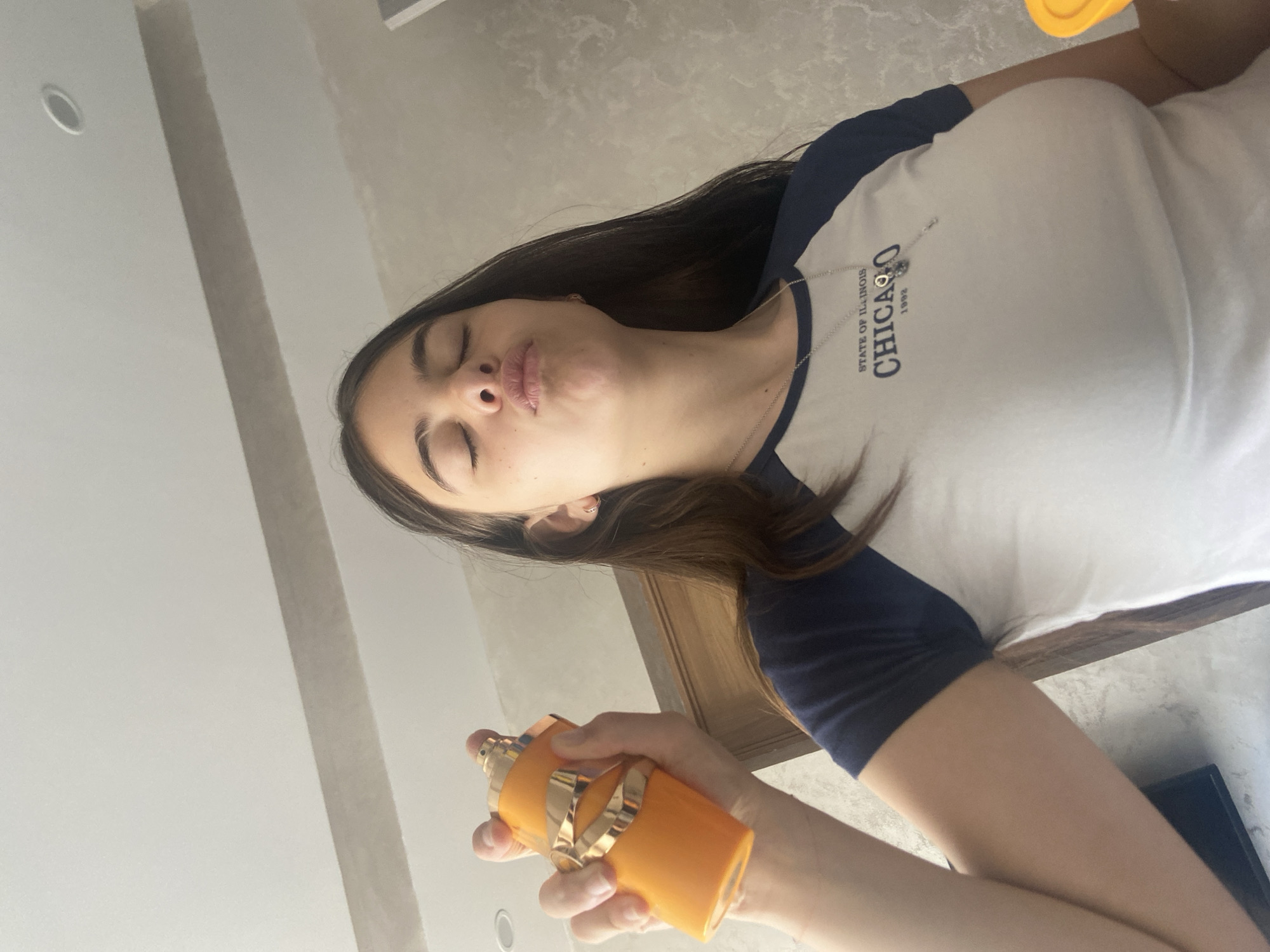 A woman is holding and spraying an orange perfume bottle. She has long brown hair and is wearing a white t-shirt with blue sleeves and the word "CHICAGO" written on it. Her eyes are closed and she is pouting. The background is blurry and shows a white ceiling and a brown wooden surface. There is no evidence in the image of Dragovishtitsa, Bulgaria.