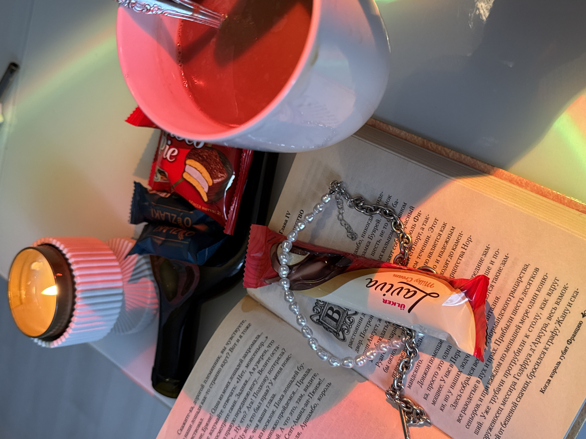 An unidentified indoor location in Unknown, Unknown presents a tabletop scene under ambient and refracted light. A lit candle in a white, ribbed holder occupies the upper left. Below and to the right, an open book displays Cyrillic text. A dark-colored bottle is partially visible behind the book. Several packaged snack items are arrayed: two dark blue foil packages (one partially legible as "O ZLAKI"), one red package with banana imagery, and one white and red package labeled "Ülker Laviva Milky Cream," resting on the book. A white ceramic mug containing a dark red liquid and a silver spoon is positioned to the right. A string of small white beads and a silver-colored metal chain are draped across the "Laviva" package and the book. Rainbow light refractions are visible across the surface, particularly in the lower left and upper right areas. No individuals are present.