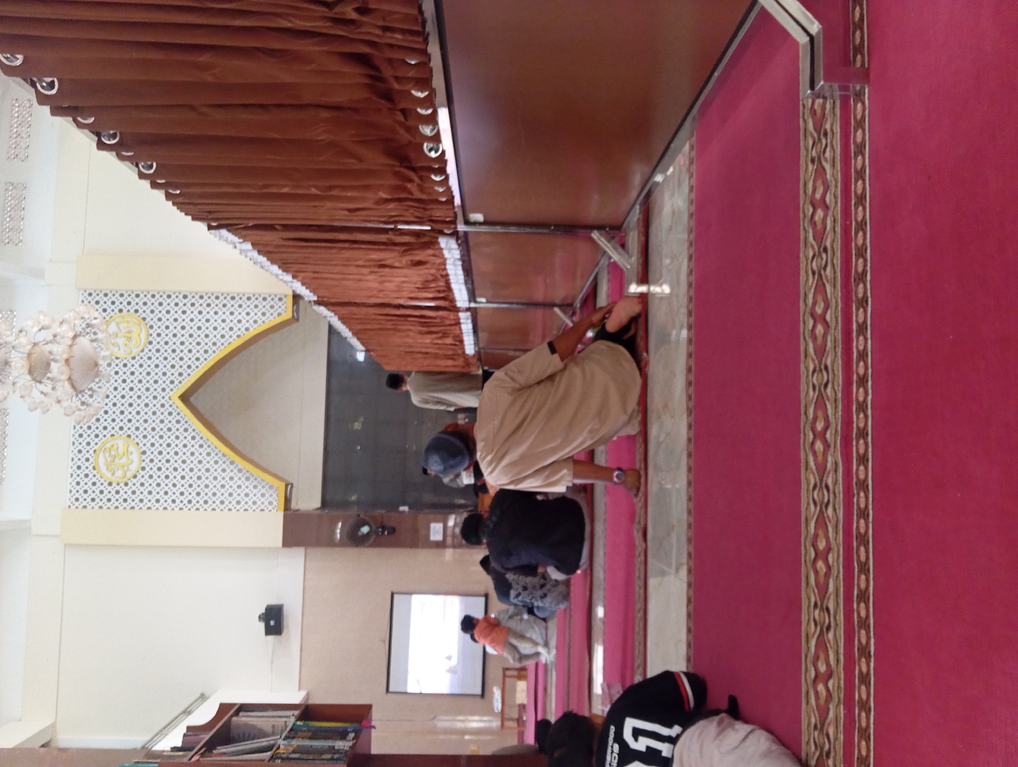 The image depicts the interior of a mosque in Moyudan, Indonesia, with a focus on the prayer hall. In the foreground, a person wearing a light brown tunic and a dark blue skullcap is seated on a red carpet, facing away from the camera. Their legs are crossed, and their right hand rests on the carpet. To the left, a child in a black jersey with white and red trim is also seated on the carpet, facing the same direction. The carpet features decorative borders with geometric patterns in red, brown, and beige.
In the mid-ground, several other individuals are seated on the carpet, some facing forward and others with their backs to the camera. One person in the center appears to be wearing a patterned garment. Further back, a projection screen is visible on the left wall, displaying an image that is difficult to discern. Adjacent to the screen, a shelf unit filled with books is present.
The background reveals the architecture of the mosque. A large, arched structure with intricate white and gold decorative panels is visible above the seating area. Brown curtains hang along the right side of the hall, partially obscuring the wall. A chandelier-like light fixture hangs from the ceiling. The walls are painted a light color, and the floor appears to be tiled in some areas. A fan is mounted on the wall in the middle ground. The overall atmosphere suggests a gathering for prayer or religious activity.