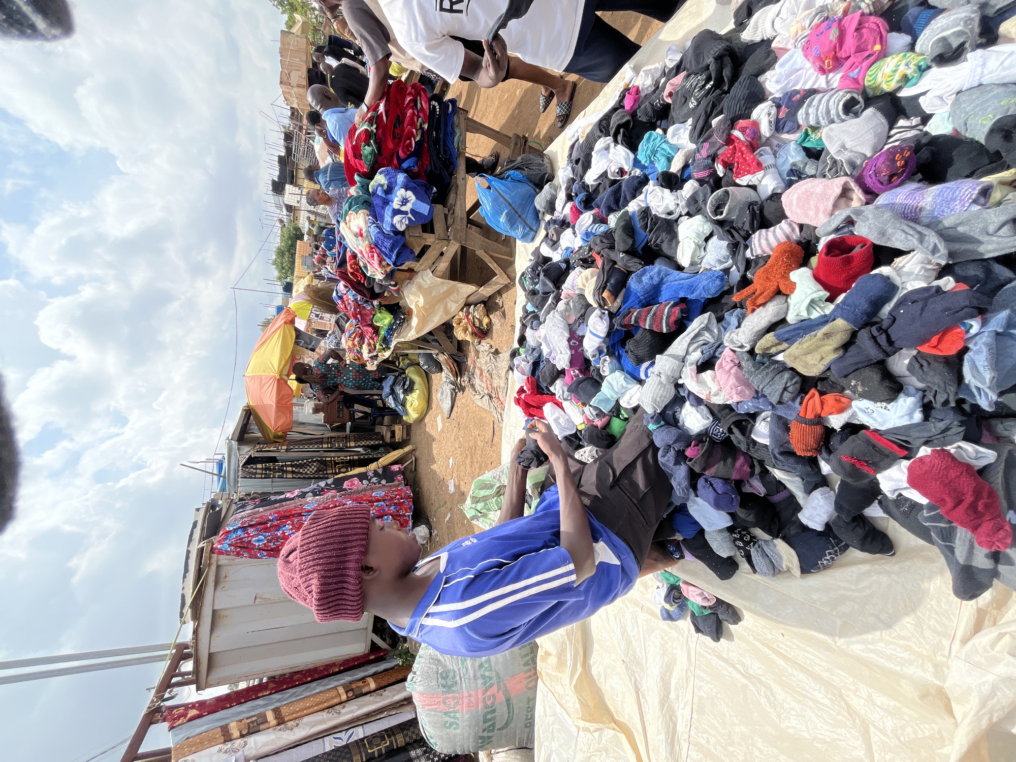 This image depicts an outdoor market scene in Gura Top, Nigeria, likely during the daytime with clear skies and abundant sunlight. The focus is on a young person wearing a maroon beanie and a blue and white striped t-shirt, who appears to be examining a large pile of socks and other assorted clothing items spread out on a light-colored tarp.
The market stall is filled with a variety of goods, including blankets folded on shelves and a large quantity of colorful fabrics stacked vertically. Behind the stall, several other individuals can be seen, some standing and others seated, amidst the bustling market environment. The overall impression is one of vibrant commerce and daily life in a Nigerian community.
The pile of clothing items is a prominent feature, showcasing a wide array of socks in different colors, patterns, and sizes, alongside other apparel such as t-shirts, hats, and possibly undergarments. The sheer volume and disarray of the clothing suggest a second-hand market or a vendor selling a large quantity of items. The individual in the foreground is engrossed in looking at the socks, perhaps making a selection or sorting through them.
The scene is rich with textural details, from the knit of the beanie and the stripes on the t-shirt to the varied textures of the fabrics and the soft sheen of the tarp. The background hints at more market stalls and structures, indicating a well-established trading area. The image conveys a sense of everyday activity, with the subjects seemingly engaged in practical tasks within their environment.