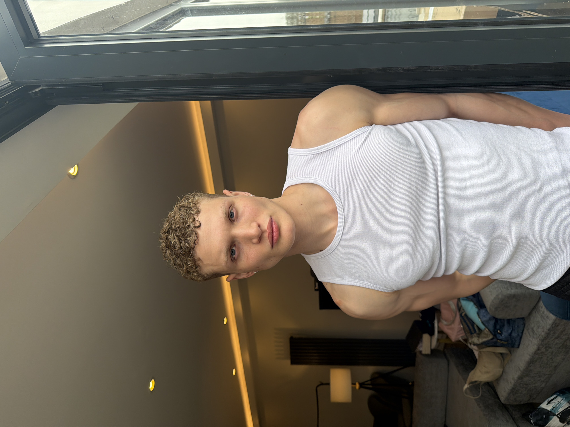 A person with short, curly blonde hair is wearing a white ribbed tank top. They are standing in front of an open window or door frame, suggesting they are indoors. The interior appears to be a modern living space with recessed lighting in the ceiling, dark furniture, and a partially visible sofa. The presence of an open window or door may suggest a view to the exterior. Given the description, it is not possible to ascertain the location in the city of London, United Kingdom.