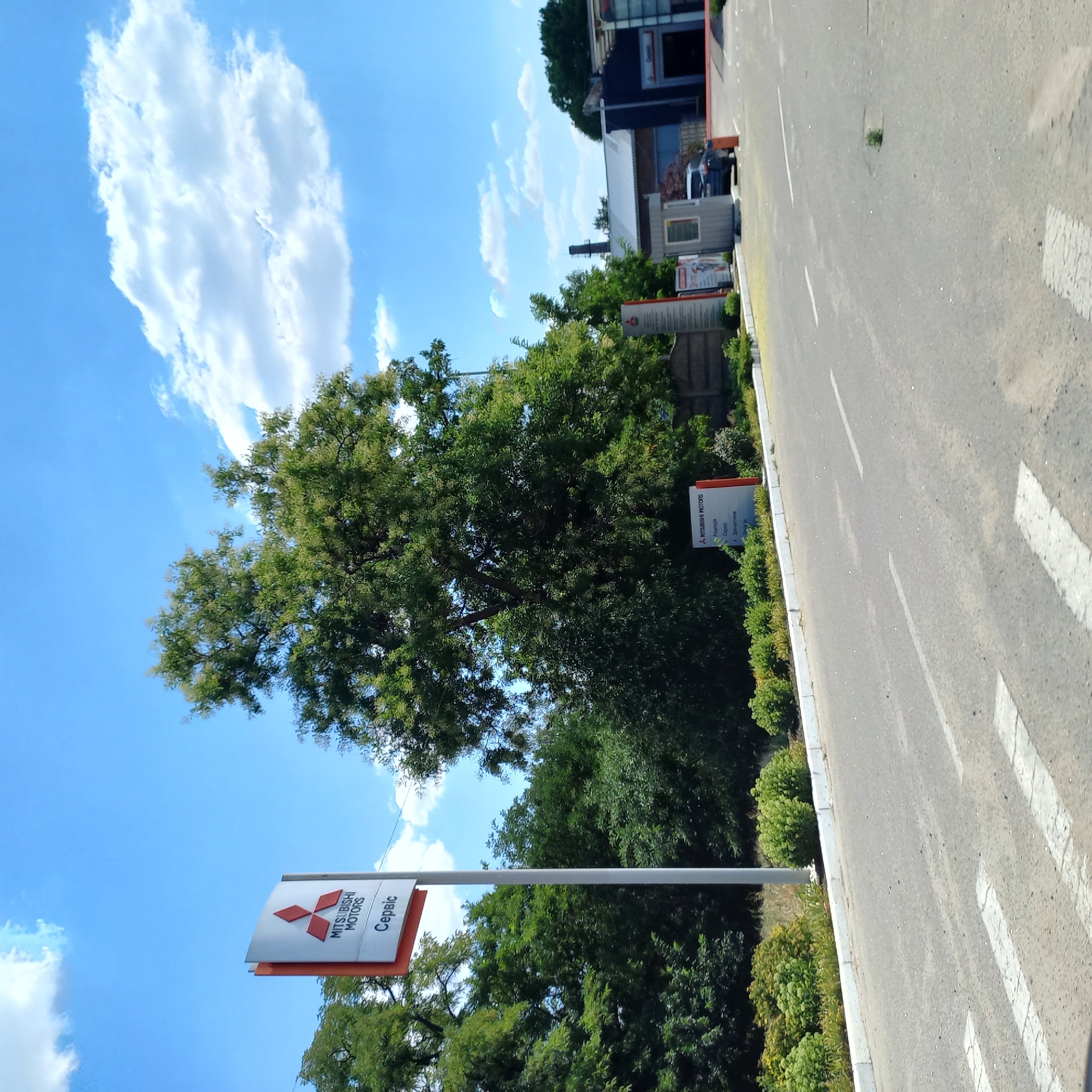 The image shows a street scene in Kropyvnytskyi, Ukraine, on a sunny day with a blue sky and scattered clouds. On the left side of the image, a tall sign for "Mitsubishi Motors Service" is visible, featuring the Mitsubishi logo and text in what appears to be Ukrainian. Behind the sign is a dense arrangement of trees and foliage.
The road in the foreground has white pedestrian crossing markings. To the right of the Mitsubishi sign, a small grey security booth or kiosk is present. Further to the right, a dark blue car is parked near a building that appears to be part of a commercial or industrial complex, possibly a car dealership or service center, with an open bay visible. The overall environment suggests an urban or suburban setting with elements of commerce and vegetation.