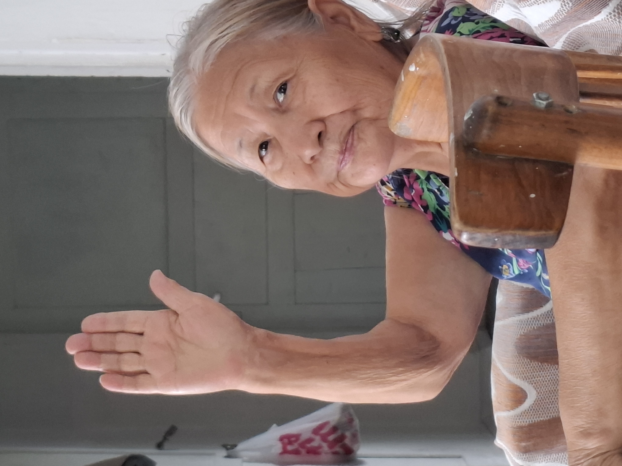 A medium shot shows an elderly woman with graying hair and light skin, looking towards the camera with a gentle smile. Her left arm is raised, with her palm facing forward in a gesture that could be interpreted as a wave or a signal. She is wearing a colorful, floral-patterned top. The woman is seated in a chair, with a wooden armrest visible in the foreground. The background is dimly lit and appears to be an indoor setting, possibly a room with a door and a plastic bag hanging on the left. The image provides no specific details about the location in Almaty, Kazakhstan.