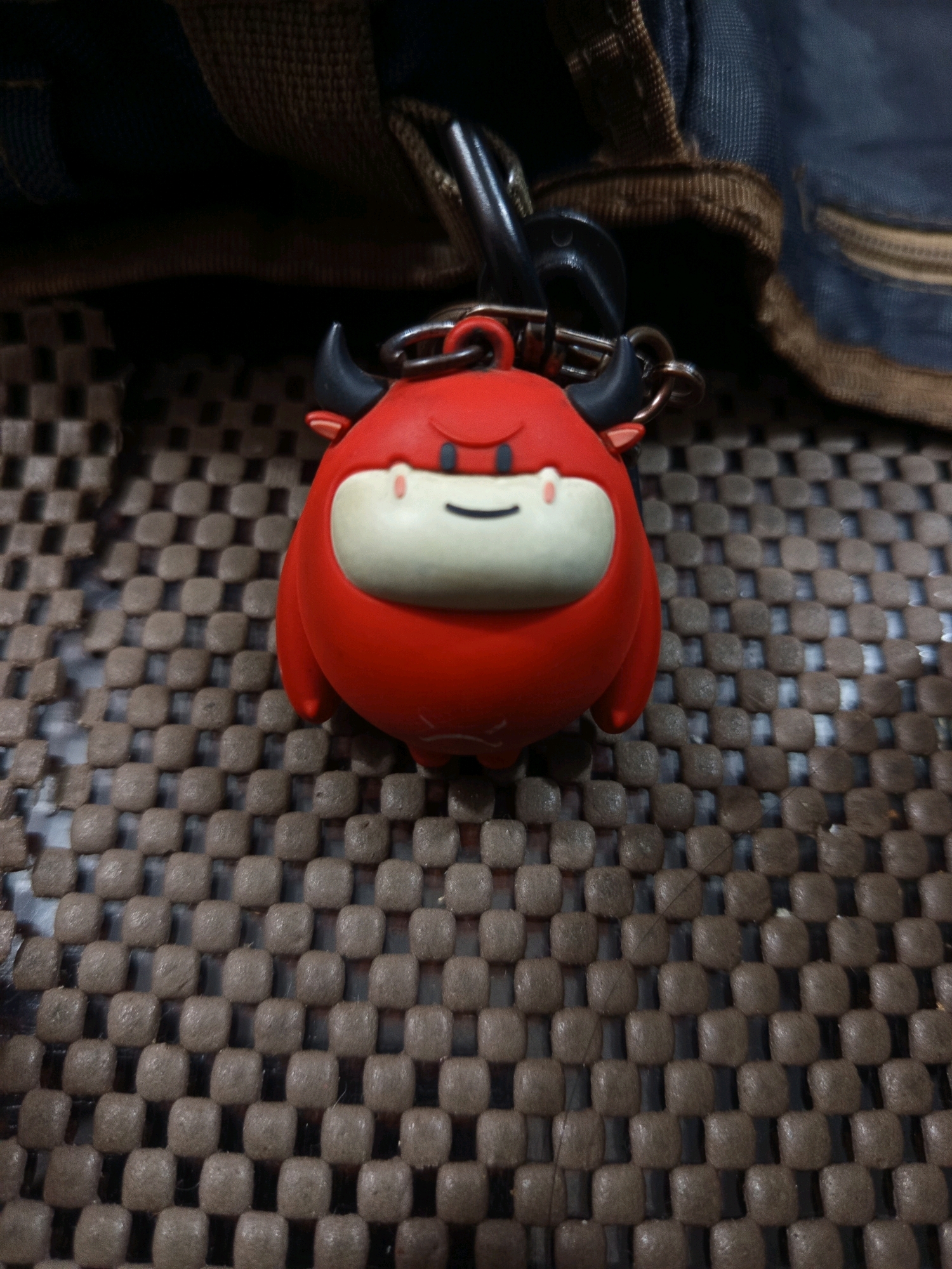 A red devil-shaped keychain with black horns and a white face with black eyes and a smiling mouth is attached to a black clasp and a silver chain. The keychain is resting on a textured brown surface. In the background, a portion of a dark blue and brown bag is visible. The image appears to be a close-up shot taken from a high angle.