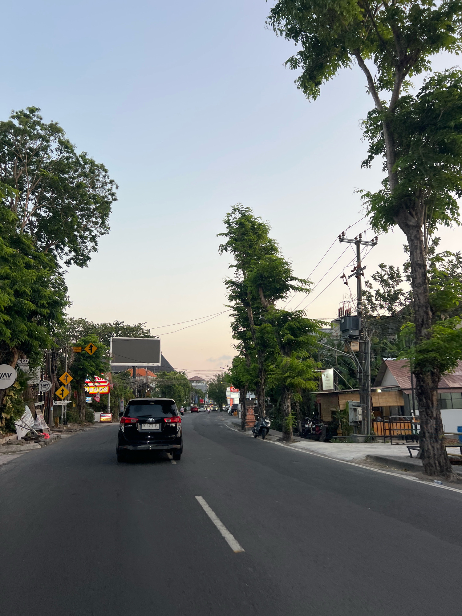 The image shows a street scene in Kuta, Indonesia, with a black SUV driving on the right side of the road. The sky is a pale blue with a hint of orange, suggesting it is either sunrise or sunset. Trees line both sides of the road, and there are buildings and shops visible on the right. A power pole with transformers and wires is also present. Several signs are visible on the left side of the road, including directional signs and advertisements. Motorcycles are parked along the sidewalk on the right. The road appears to be paved and has a white dividing line. The overall impression is of a typical street scene in a tropical urban environment.
