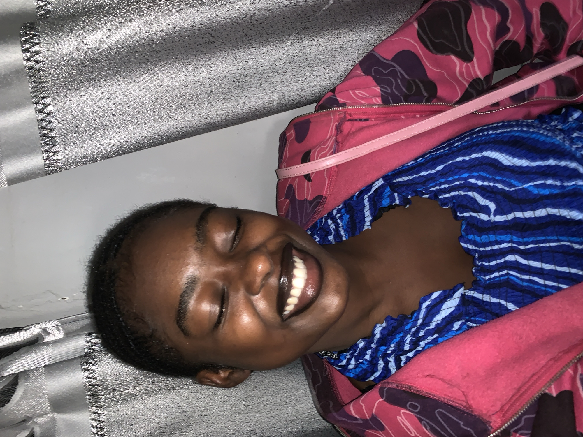 The image depicts a woman lying down. She has dark skin, short black hair, and is smiling widely, revealing her teeth. She is wearing a blue and white patterned top, layered under a pink zippered hoodie with a darker pink and purple pattern. A light pink strap is visible across her chest. The background consists of a textured silver or gray surface. It's impossible to verify the location as Rafin Par, Nigeria without further information.