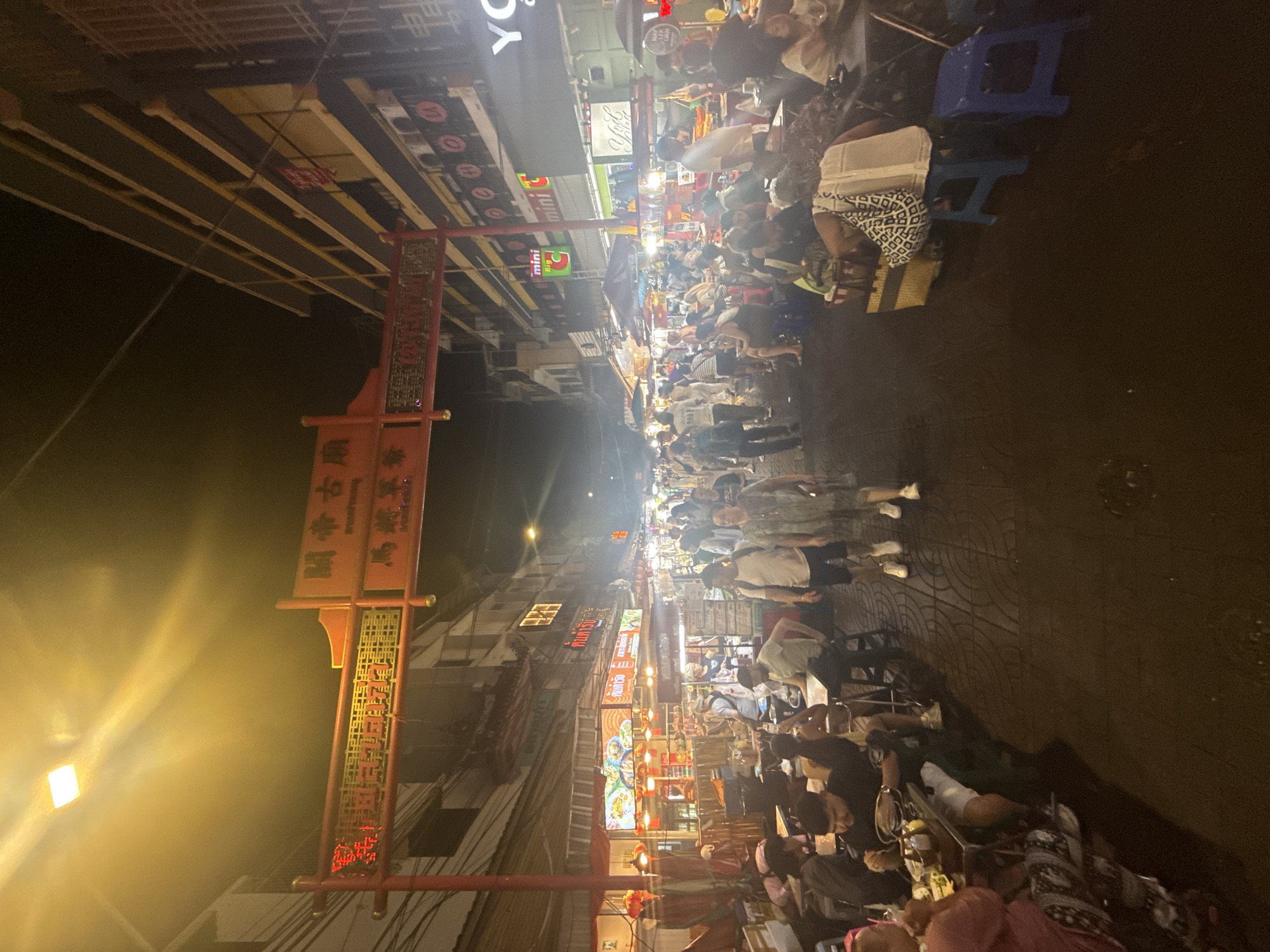 This nighttime image captures a bustling street food market in Yaowarat, Bangkok's Chinatown, Thailand. The central feature is a red Chinese-style archway spanning the street, displaying "เยาวราช" (Yaowarat) in Thai script on the left and Chinese characters, including "古關帝廟" (Guandi Temple), on the right.
The street, appearing to be pedestrianized, is densely populated with individuals of various ages, primarily engaged in eating, drinking, socializing, and walking. Both sides of the street are lined with multi-story buildings, their ground floors occupied by numerous street food stalls and small eateries. Patrons are seated at small, simple tables with plastic stools, consuming food and beverages. Vendors are visible preparing and serving food from mobile carts and fixed stalls.
The scene is heavily illuminated by a multitude of artificial light sources, primarily warm yellow and orange, emanating from overhead streetlights, shop signs, and individual food stalls, casting a glow in the upper sky. Visible signage includes Thai and Chinese scripts, with an English "mini Big C" logo and a partially visible "YO..." sign on the right. Electrical wires are strung overhead between buildings. The ground is paved with patterned tiles. The overall environment is lively and crowded, indicative of a popular urban night market.