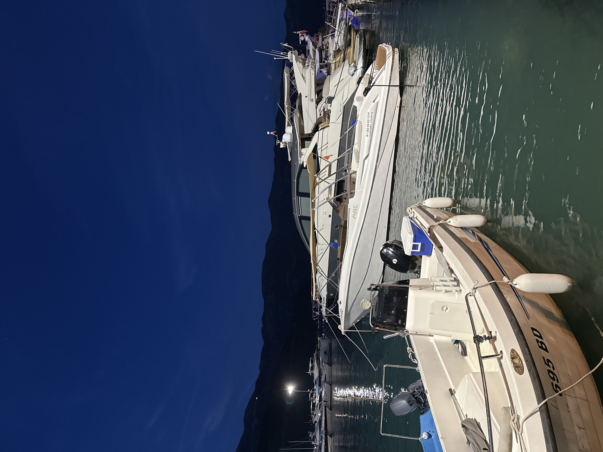 The image depicts a harbor scene at night in Budva, Montenegro. Several yachts and smaller boats are docked. In the foreground, a white boat with the registration "695 BD" is visible, equipped with two outboard motors. In the mid-ground, a white yacht with the text "ARC" and contact information "+382 69 821 310 | adriatic_cruising" is prominently featured. Further in the background, more yachts of varying sizes are moored, their reflections shimmering on the dark water. The sky is a deep blue, transitioning to black towards the horizon, with distant mountains silhouetted against the night sky. A single bright light illuminates the scene from a lamppost on the left side of the image. The water is calm, with gentle ripples reflecting the ambient light. The overall atmosphere is serene and tranquil.