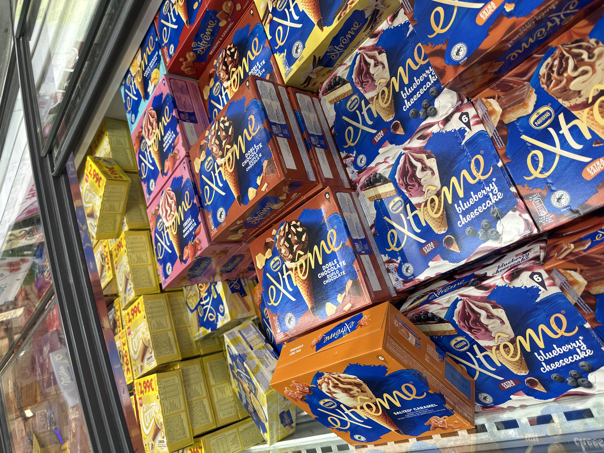 The image captures a vibrant display of ice cream cones in a supermarket freezer aisle. The scene is dominated by numerous boxes of "Xtreme" brand ice creams by Nestlé, stacked neatly on shelves inside a glass-fronted freezer.
The "Xtreme" boxes come in various colorful designs, indicating different flavors. Prominently visible are "blueberry cheesecake" (blue packaging), "DOBLE CHOCOLATE / DUPLO CHOCOLATE" (brown packaging), and "SALTED CARAMEL" (orange packaging). Other discernible flavors include "PEANUTS & CARAMEL", "FRESA / MORANGO & SABOR NATA" (strawberry), and "DOBLE VAINILLA DUPLO BAUNILHA" (double vanilla). Many boxes indicate "6" cones per pack and feature an "EXTRA SAUCE" label, along with a cocoa bean symbol.
On the left side, behind the glass door, several yellow boxes of "Cornetto" brand ice creams are visible, each marked with a price of "2,99€" and the flavor "LEMON". Some of these boxes also feature a "SUPER POUCO" or "SUPER POCO" sticker.
The glass freezer door reflects ambient store lighting and other products, including a faint "Milka" logo. The boxes are arranged densely, suggesting a well-stocked freezer. The overall impression is a colorful and appealing display of frozen treats, likely aimed at shoppers. The visible pricing in Euros suggests the location is in a European country.