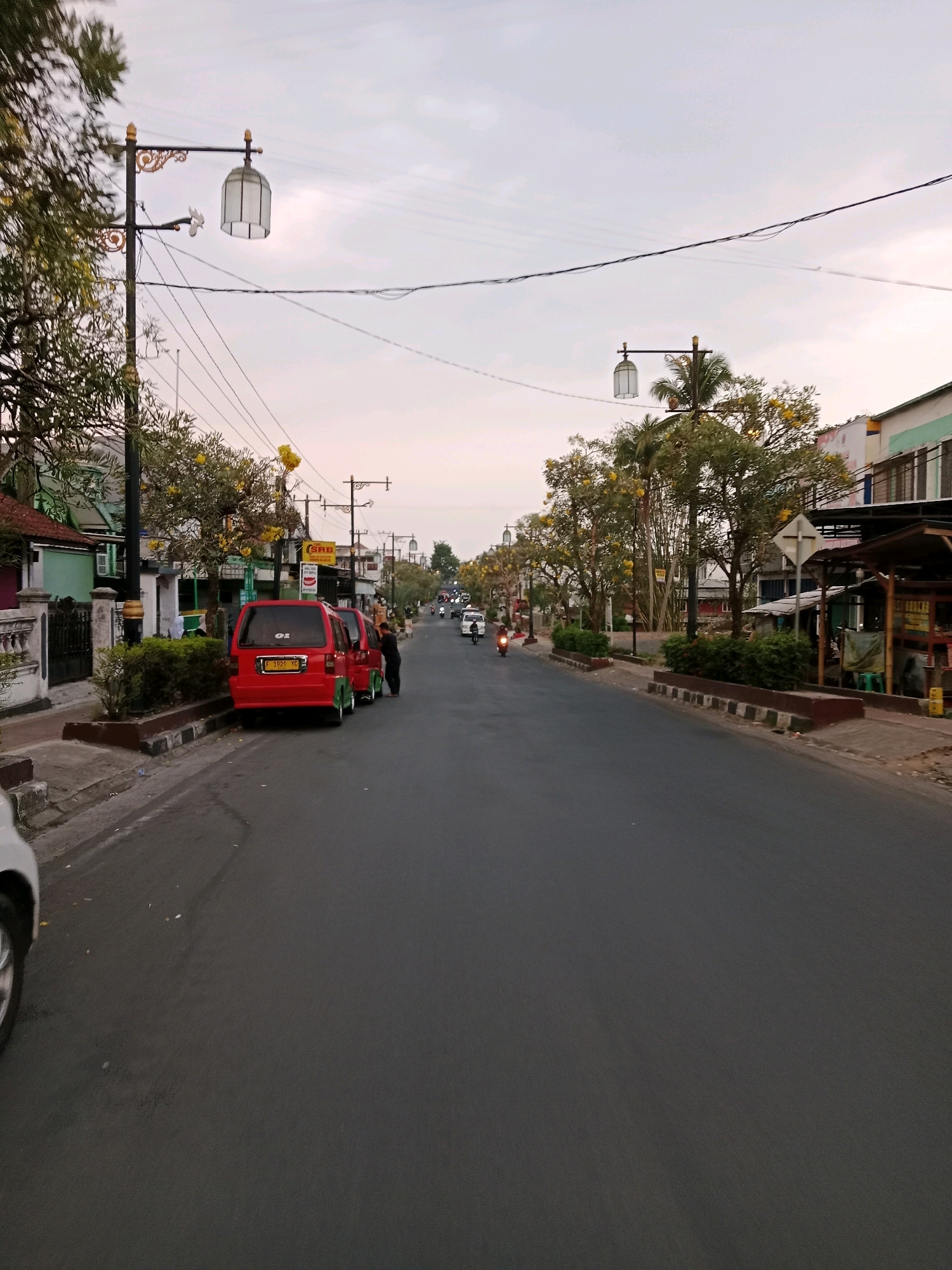 This is a street-level, medium shot image of a road in Cianjur, Indonesia. The sky is overcast with a light, hazy glow, suggesting either early morning or late afternoon. The road is asphalt and appears to be in moderate condition, with some visible texture and a slight sheen.
On the left side of the road, a sidewalk runs parallel, bordered by a low concrete wall and a strip of vegetation. A white car is partially visible on the far left, its front end cut off by the frame. Next to the sidewalk, a red minivan is parked, facing away from the camera, with its rear visible. Two people are standing near the back of the minivan, one appearing to interact with the vehicle. A second red minivan is parked behind the first, also facing away.
Further down the street, trees with yellow flowers line the sidewalk, interspersed with decorative lampposts that feature ornate gold accents and white, cage-like lanterns. Power lines are strung overhead, crossing the sky. Buildings with various architectural styles and colors line the right side of the road, some appearing to be shops or businesses, with awnings and signage visible. One sign prominently displays "SRB".
Several vehicles, including motorcycles and other cars, are present on the road, moving in the distance. The overall atmosphere is that of a quiet urban street, possibly in a residential or commercial area, during a transitional time of day.
