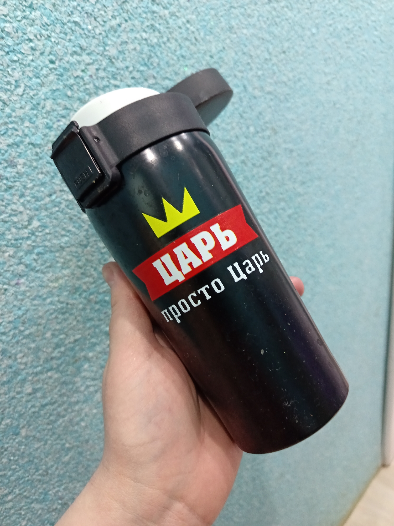 A close-up shot captures a person's hand holding a black thermos or travel mug indoors, in Osa, Russia. The thermos is the primary subject, held slightly to the right of the frame, with a light blue, textured wall serving as the background.
The black thermos features a distinctive design: a bright yellow crown graphic is emblazoned on its side. Beneath the crown, a red banner extends across the mug. Visible white Cyrillic text is present on the design: "ЦАРЬ" (TSAR) is written within the yellow crown, and "просто Царь" (simply a Tsar) appears on the red banner below it. The lid of the thermos is black with a white inner section, and a small black clasp on the side clearly displays the word "PUSH". The hand holding the mug shows visible skin texture. The background wall has a coarse, granular texture and is uniformly light blue. A narrow vertical strip of a white door frame is visible on the far right side of the image, suggesting an indoor setting. No specific time of day or emotions are discernible.