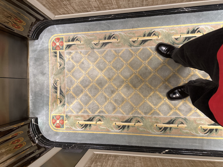 An overhead view captures the lower extremities of a person standing inside an elevator, likely within The Langham London hotel in Marylebone, London, United Kingdom. The individual is wearing polished black leather loafers and dark trousers; a red garment is visible across the bottom of the frame. The elevator floor is covered by an ornate, light grey carpet featuring a central yellow diamond lattice pattern. A border surrounds this pattern, depicting stylized green foliage and prominent red rose emblems in each corner. The elevator's interior includes dark, polished, multi-layered corner columns, possibly of marble, and reflective panels above.