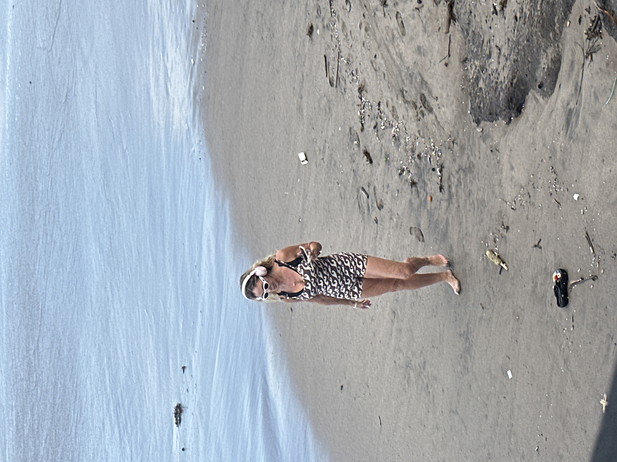 A woman with blonde hair, wearing white-rimmed sunglasses and pink headphones, walks along the sandy shore of a beach. She is wearing a patterned one-piece swimsuit with a design of brown and white wavy lines. She holds a white object, possibly a phone, in her right hand. The ocean is calm and blue to her left, and the beach sand is wet in places. Scattered on the sand are small shells, pieces of driftwood, and what appear to be footprints. In the foreground, a black sandal with colorful embellishments lies on the sand. The scene appears to be in North Kuta, Indonesia, based on the environmental context.