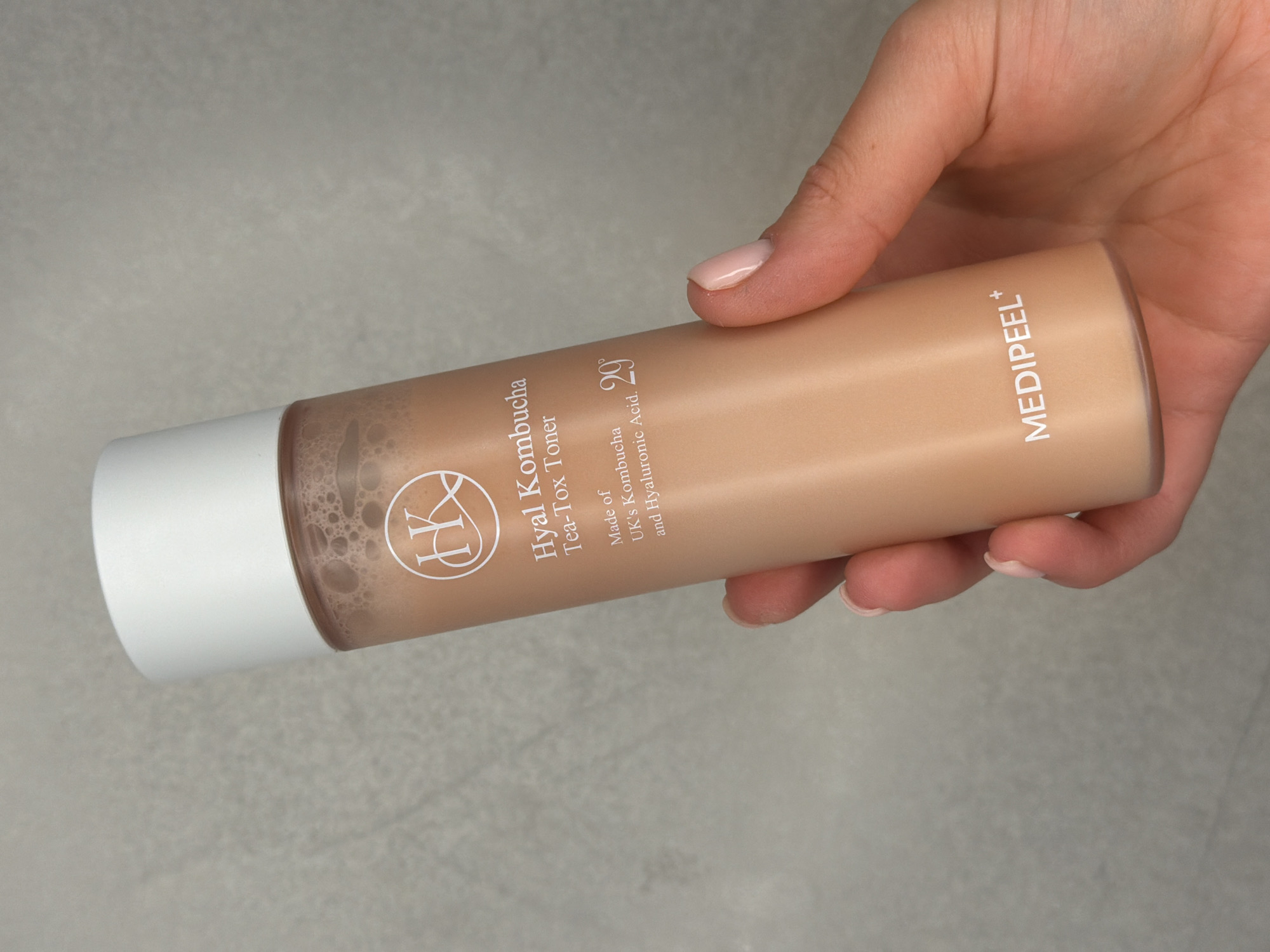 A light-skinned hand, with neatly manicured, pale pink nails, holds a cylindrical cosmetic bottle horizontally against a soft, light gray, textured background. The bottle, primarily a warm beige or light brown color, has a clean white cap on its left end. Inside, a light brown liquid is visible, with numerous small bubbles concentrated near the cap, suggesting it might have been recently shaken.
The bottle features white lettering and a logo. A stylized "HK" monogram is prominent, followed by the product name: "Hyal Kombucha Tea-Tox Toner." Below this, text reads, "Made of UK's Kombucha and Hyaluronic Acid." The number "29" is also printed, likely indicating a percentage or concentration of an ingredient. Towards the right side of the bottle, the brand name "MEDIPEEL+" is displayed vertically.
The image is a clear, focused product shot, likely taken indoors to highlight the cosmetic toner. The overall presentation is clean and highlights the product's details. This scene is situated in Sofiivska Borshchahivka, Ukraine.