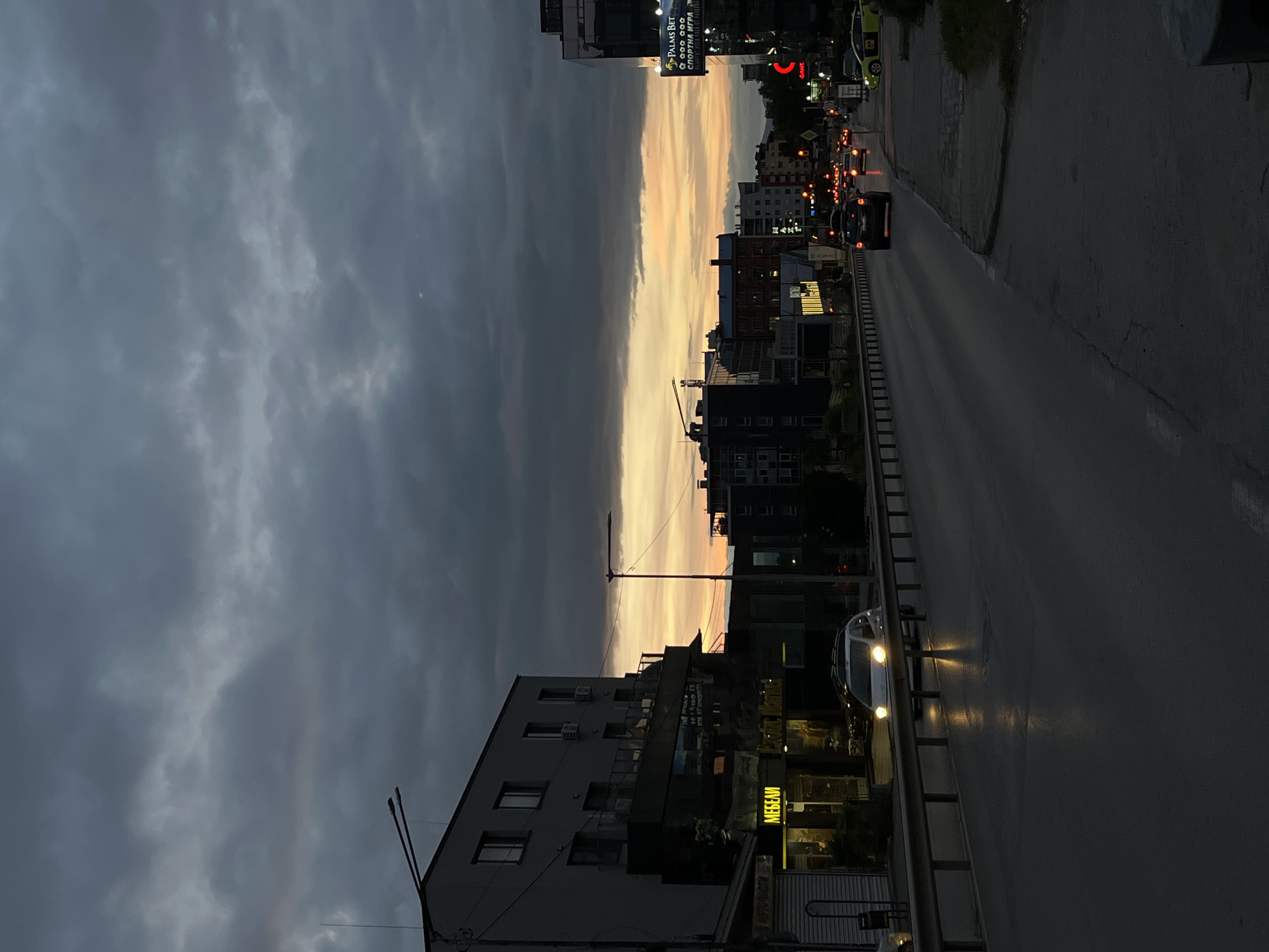 The image depicts a street scene in Sofia, Bulgaria, at dusk. The sky is filled with heavy, dark clouds, with a band of orange and yellow light visible on the horizon, suggesting the setting sun. The street is wet, reflecting the headlights of cars and streetlights.
On the left side of the street, there is a multi-story building with a dark facade. A sign with yellow lettering that reads "МЕБЕЛИ" (Mebeli, meaning furniture in Bulgarian) is prominently displayed on the lower level of the building. Adjacent to this, there are other businesses with illuminated signs.
The road itself is a multi-lane thoroughfare with a metal guardrail separating the traffic lanes from the sidewalk or adjacent property. Several cars are visible on the road. One white car with its headlights on is approaching the viewer. Further down the road, more cars with their taillights illuminated are moving away.
On the right side of the street, another multi-story building stands, featuring large billboards. One billboard advertises "PALMS BET" and "СПОРТНА ИГРА" (Sportna Igra, meaning sports game). Further down, a yellow taxi is parked or moving slowly. The overall impression is a busy urban environment transitioning into nighttime.