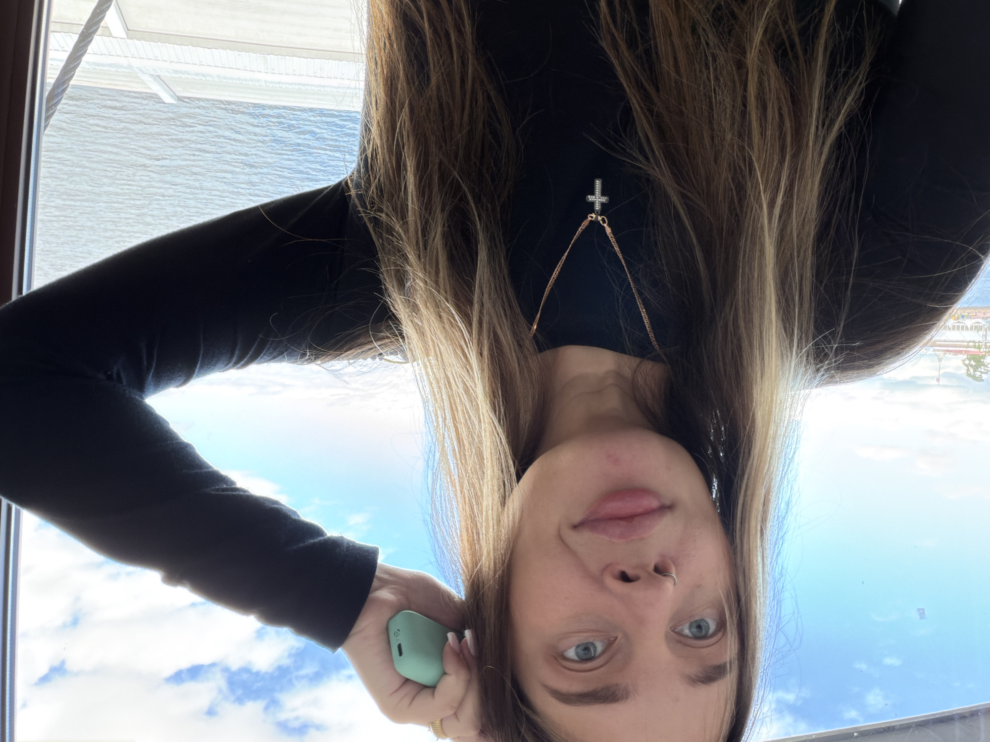 The media file is a close-up, upside-down selfie of a young person with long, brown hair with blonde highlights. They are wearing a black, long-sleeved shirt and a delicate cross necklace with small gems. The background is a bright blue sky with some white clouds, suggesting it is daytime. A body of water is visible in the upper left portion of the image, and what appears to be a building or structure with a metal railing is also present. The person is holding a small, light green object, possibly a mobile phone or a vape, in their left hand. Their right hand is near their face, and they are wearing a ring on their right ring finger. The person has a nose ring and light blue eyes. The overall mood appears casual and relaxed. No visible text is present.