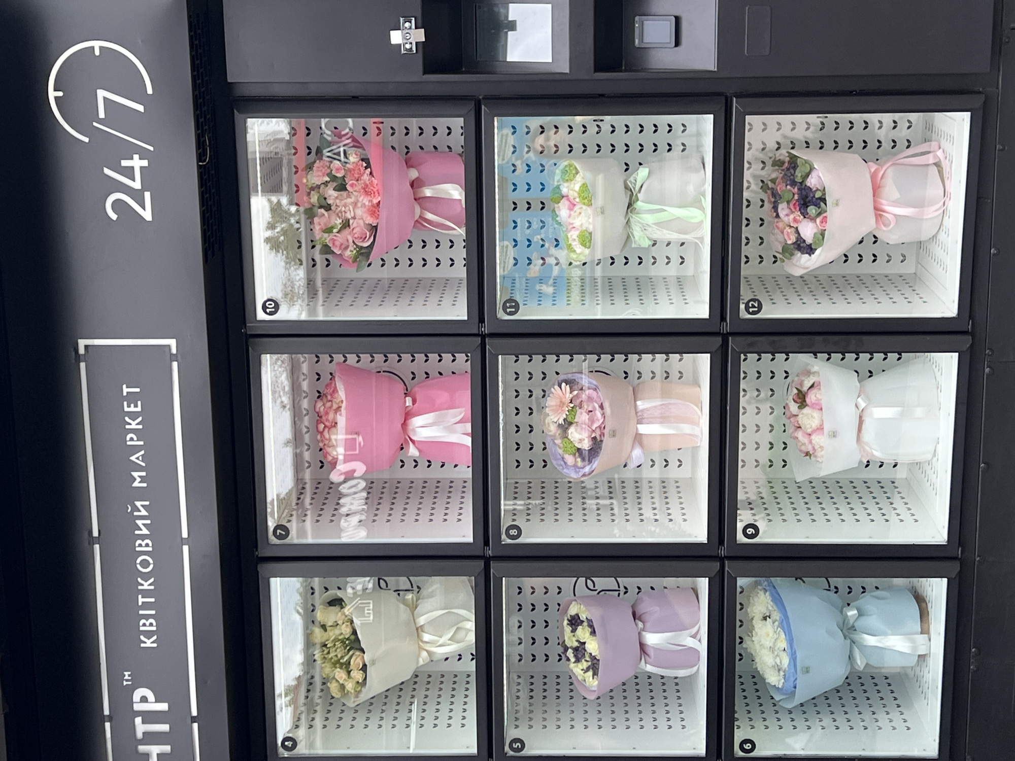 The image captures a vending machine selling bouquets of flowers, likely in a public space in Ternopil, Ukraine. The vending machine is black and features a "24/7" logo, indicating around-the-clock availability. The logo includes a clock icon. Next to the logo is some white vertical text written in Ukrainian. Each bouquet is displayed in a separate, numbered compartment, ranging from 4 to 12, likely indicating different price points or flower types. Each compartment is lit, showcasing the different bouquets: some are wrapped in pink paper with white ribbon, others are wrapped in white paper with green ribbon, and others are wrapped in white or blue paper with white ribbon. The bouquets feature a variety of flowers, including roses and carnations. Above the flower compartments are control panels and payment slots. The general environment appears clean.