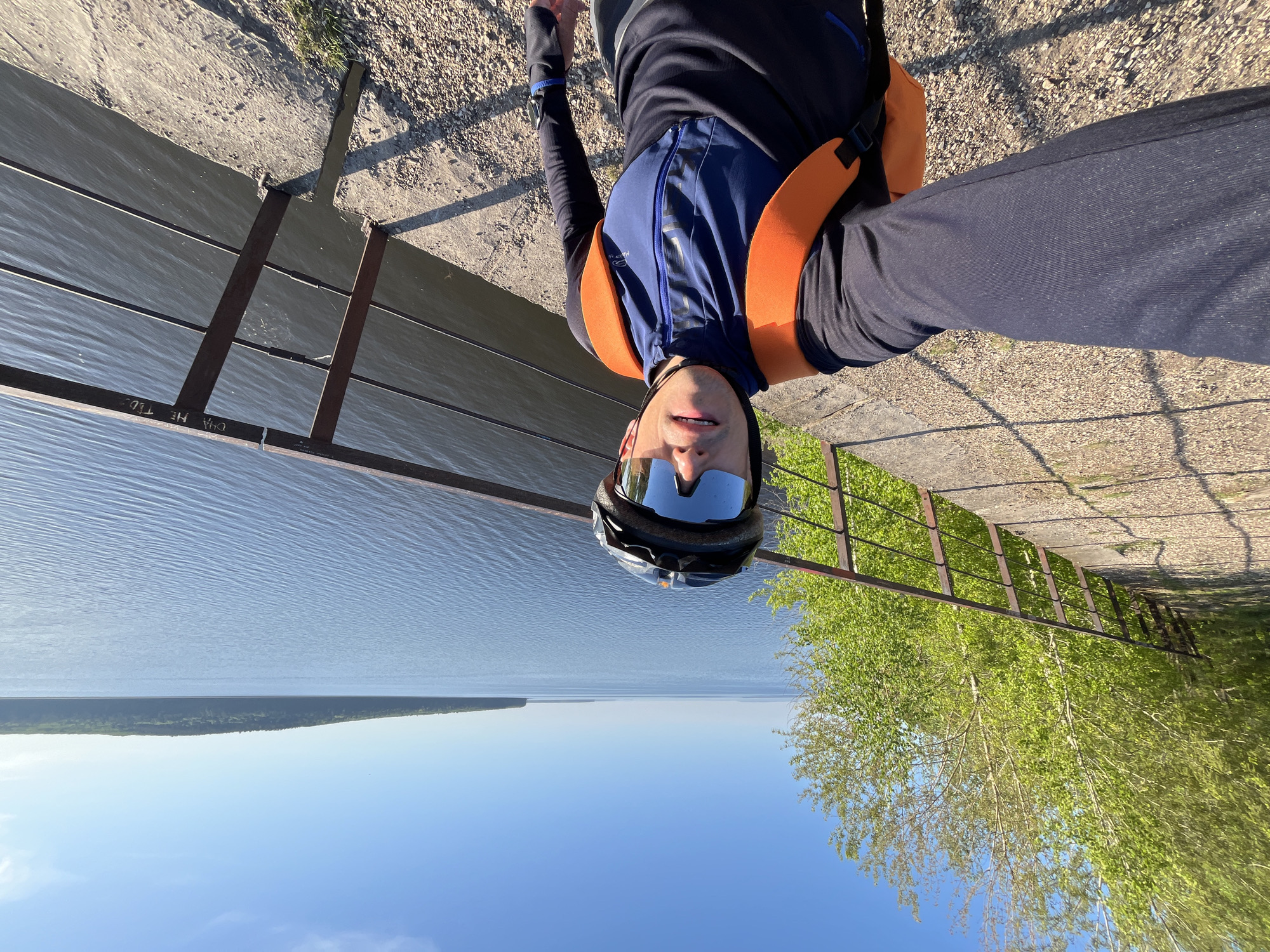 A selfie photograph shows a person wearing a helmet and an orange life vest, oriented upside down relative to the landscape behind them. The person is standing near a body of water, presumably the Kama River, based on the location being identified as Chaikovsky, Russia. A metal fence separates the person from the water. Behind the fence is a view of the river, a distant shoreline, and the sky. There are green trees visible on the other side of the fence. The person is wearing dark clothing. The ground beneath the person's feet appears to be a textured surface.