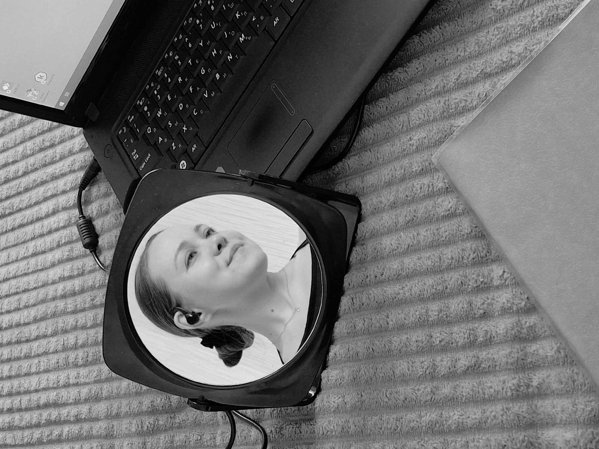 The image, in grayscale, depicts a scene with a laptop, a mirror reflecting a woman's face, and a textured surface, likely a towel. The laptop is open, with the keyboard and part of the screen visible. A round mirror, possibly for makeup or grooming, rests on the towel near the laptop. The mirror reflects the face of a young woman with her hair pulled back. The towel is light-colored, with a raised, corded texture. Given the laptop keyboard with Cyrillic lettering, it is likely in a Russian-speaking country, and given the prompt, could potentially be Chaikovsky, Russia.