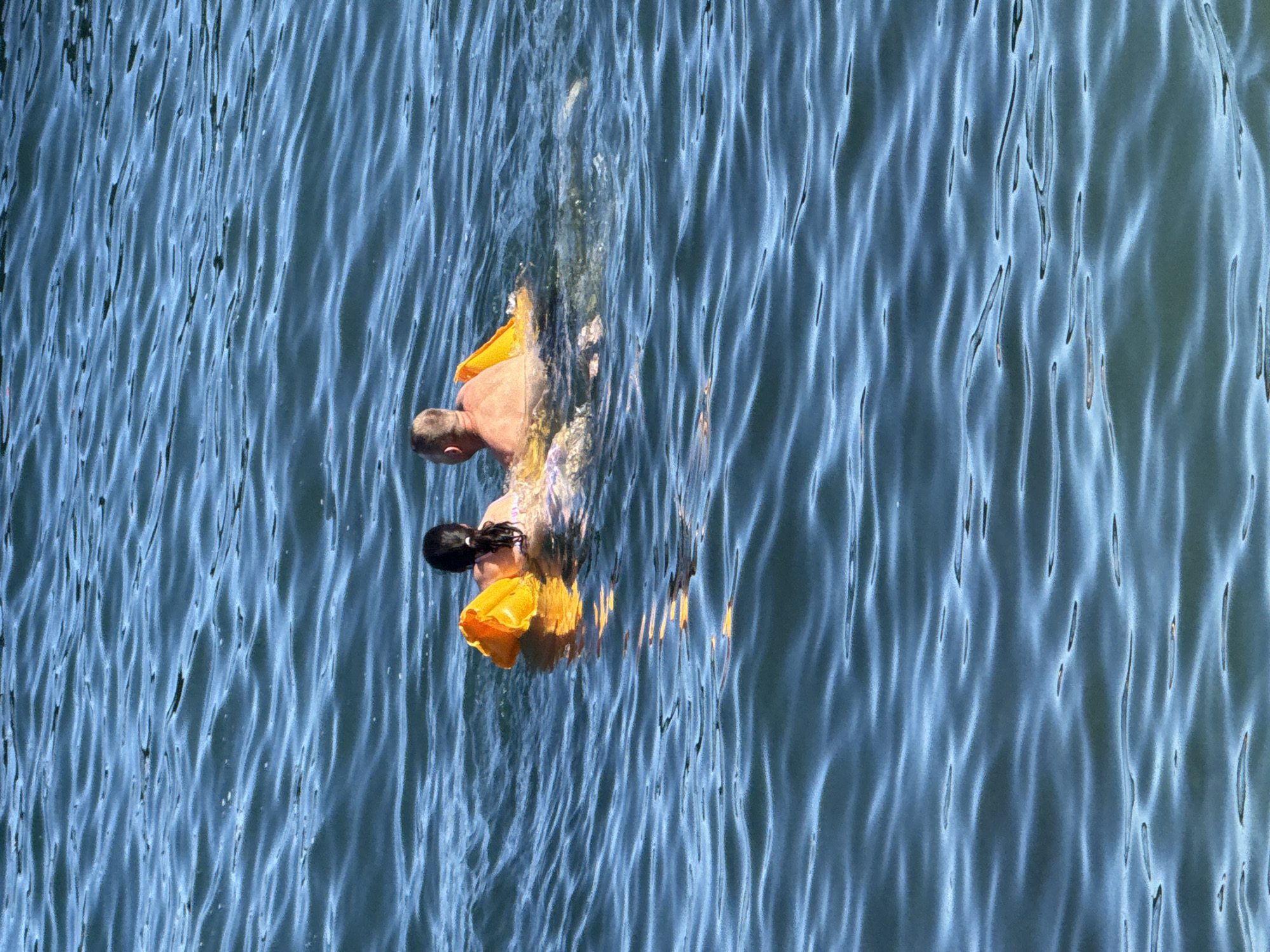 In the blue waters of the Black Sea off the coast of Partenit, Ukraine, a man and a woman are swimming. They are partially submerged in the water, with the man on the right and the woman on the left. Both are facing away from the camera. They appear to be using an inflatable yellow float, which is partially visible between them. The water is calm with gentle ripples. The sky is not visible in the frame.