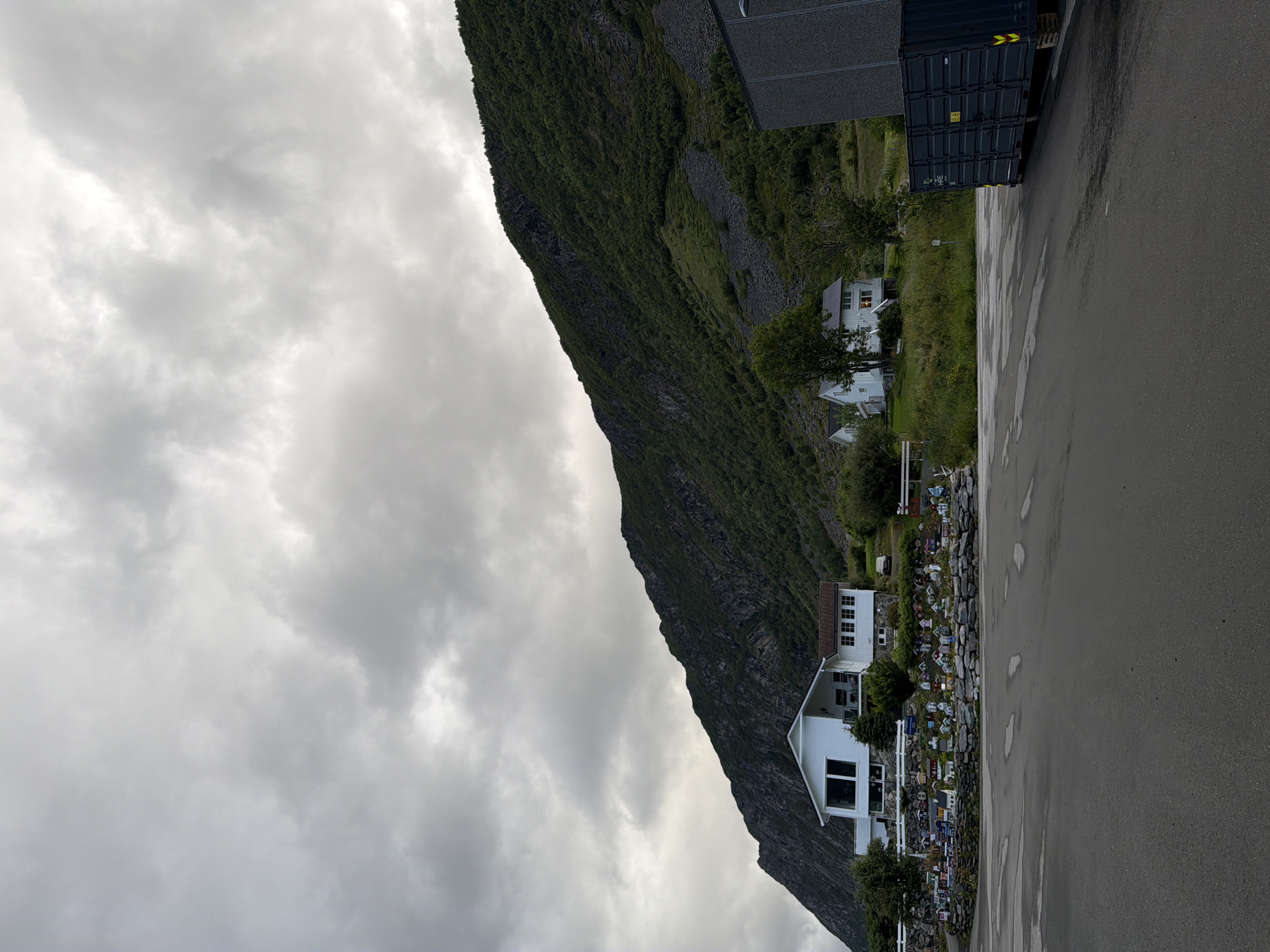 This is a photograph taken on a cloudy day, with a dramatic mountain slope dominating the background. The mountain is covered in dense green vegetation, with patches of exposed rock. In the foreground, there is a large, dark grey, industrial dumpster. To the left of the dumpster, there are two white houses. One house is larger and more modern with large windows, while the other is smaller and more traditional. In front of the houses, there is a collection of miniature houses, arranged along a low stone wall. The ground in the foreground is asphalt, wet and reflective, with puddles suggesting recent rain. The overall scene evokes a sense of rural or semi-industrial coastal Norway, with the specific location likely being in or near Harstad.