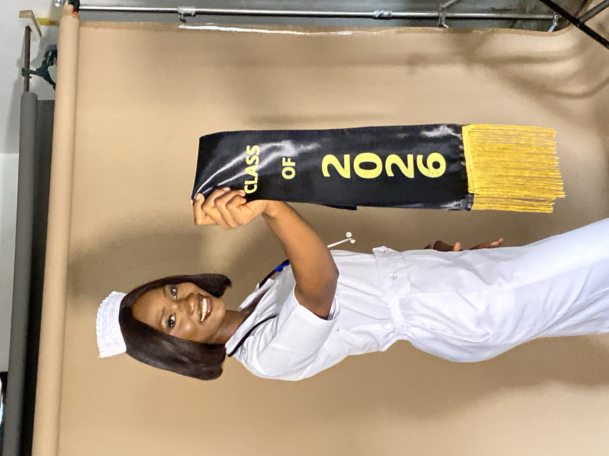 A woman wearing a white nurse's uniform and a nurse's cap is posing for a photograph in front of a beige backdrop. She is holding up a black graduation sash with "CLASS OF" and "2026" printed in yellow letters. The sash also has yellow fringe. The location is a photo studio, likely in Gura Top, Nigeria.