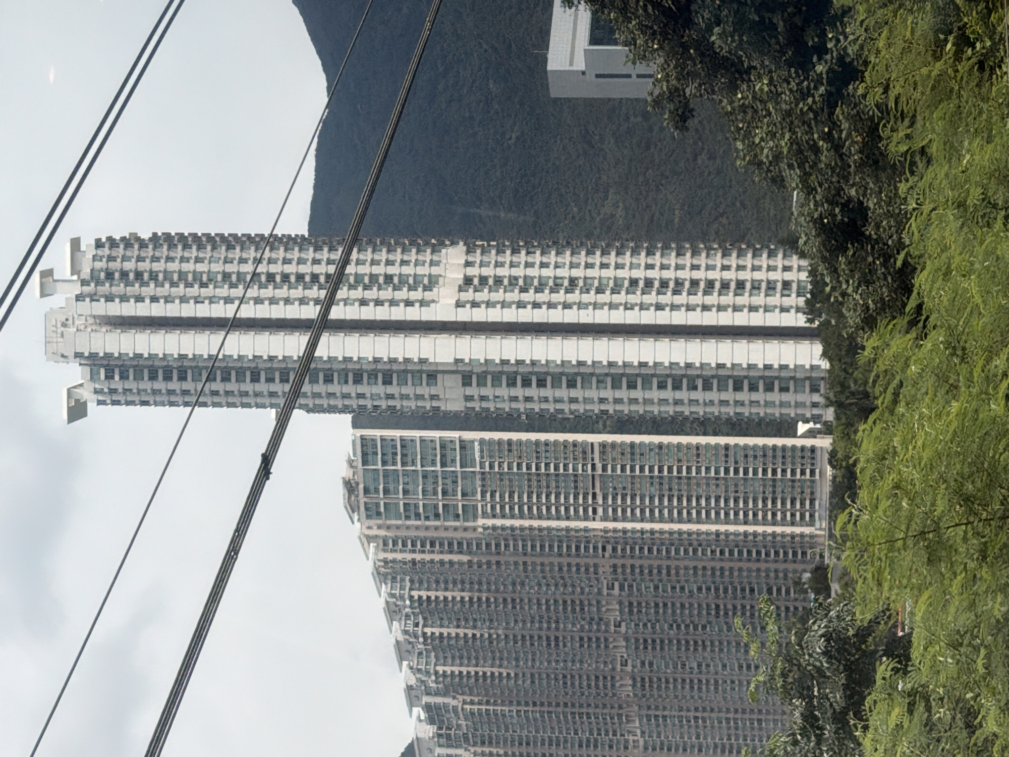 A medium shot captures a panoramic view of a dense urban landscape in Hong Kong, China, characterized by towering residential skyscrapers and lush green hills. The dominant features are several tall, modern apartment buildings, their facades a mosaic of glass windows and white architectural elements. The buildings vary in height and design, with one particularly slender, white skyscraper standing out in the center, adorned with unique angular roof structures. To the left, a cluster of even taller, densely packed residential towers forms a wall of urban development, their repetitive window patterns suggesting a high-density living environment. In the background, a steep, verdant mountain slope rises, its dark green foliage contrasting with the bright urban structures. A modern, white rectangular building with dark windows is visible on the right side, partially obscured by dense trees. In the foreground, a thick canopy of vibrant green trees and shrubs creates a natural foreground, with the foliage appearing lush and well-maintained, indicating a humid, possibly tropical climate. Several dark, horizontal cables stretch across the upper portion of the frame, suggesting the presence of a cable car system or power lines, adding a layer of infrastructure to the scene. The sky above is overcast, with a pale, diffused light illuminating the cityscape. There are no people or animals visible in the image, focusing the viewer's attention on the architectural and natural elements of the environment.