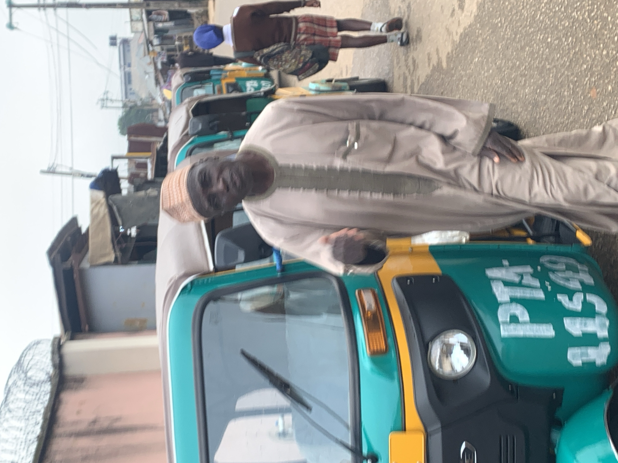 A man in traditional light brown attire and a patterned cap stands next to a teal and yellow auto-rickshaw, partially obscured by the vehicle. His right hand is placed on his hip. In the background, another auto-rickshaw is visible, and a person wearing a blue headwrap, brown sweater, and plaid shorts walks away from the camera. The scene appears to be on a paved street with buildings and power lines in the background, suggesting an urban setting in Jos, Nigeria. The weather is overcast. The auto-rickshaw has "PTA 11549" written on its side in white lettering.