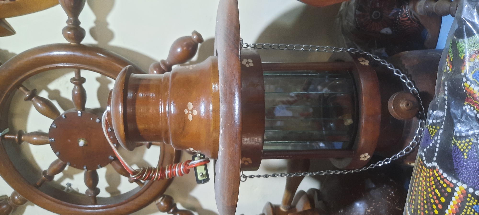 The image depicts a close-up, vertical view of a decorative wooden lantern with glass panels, suspended by chains. The lantern has a dark brown finish and is adorned with small, lighter-colored floral motifs. It rests on a flat, circular wooden base. Above the lantern, a portion of a large wooden ship's wheel is visible, also dark brown and intricately carved. Wires, one red and white, are loosely coiled and attached to the top of the lantern, with a small battery-like object visible near the coiled wires.
The background is a plain, light-colored wall. In the foreground, partially visible, is a colorful, patterned fabric or wrap, possibly an Indonesian batik or textile, with intricate dot patterns in yellow, orange, purple, and white on a dark background. To the right of the lantern, a portion of another carved wooden object, possibly a decorative mask or figurine, is visible. The scene appears to be an indoor display, likely in a shop or home, showcasing decorative items.
Based on the available information, this scene is likely located in the city of Blitar, Indonesia, given the context of the provided image and the commonality of such decorative items and artistic crafts in the region. The precise location within Blitar is not discernible from the image itself. No people are visible in the image, and no actions or interactions are depicted. The environment is static and appears to be a curated display of artisanal objects.