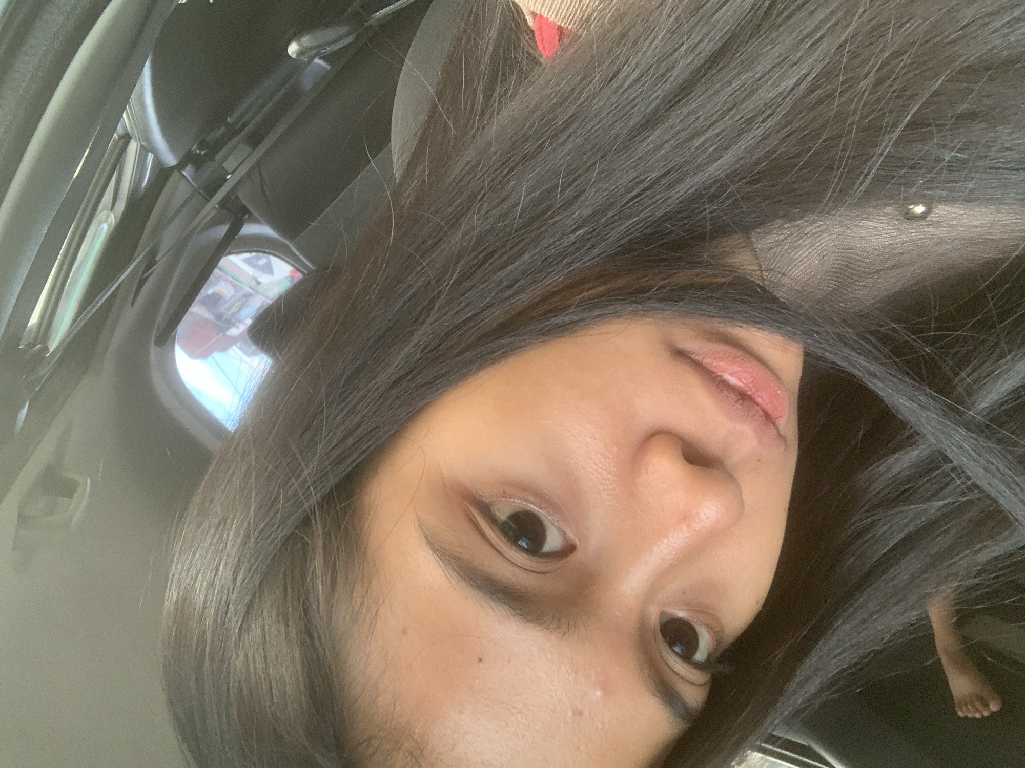A close-up, slightly tilted, selfie shows a woman with long, dark hair. She appears to be inside a vehicle, possibly a car, as indicated by the interior elements visible in the background and the reflection in the window. A foot is visible in the lower right quadrant. The context suggests this may be Samarinda, Indonesia, but there are no directly visible identifiers to explicitly confirm the exact location.