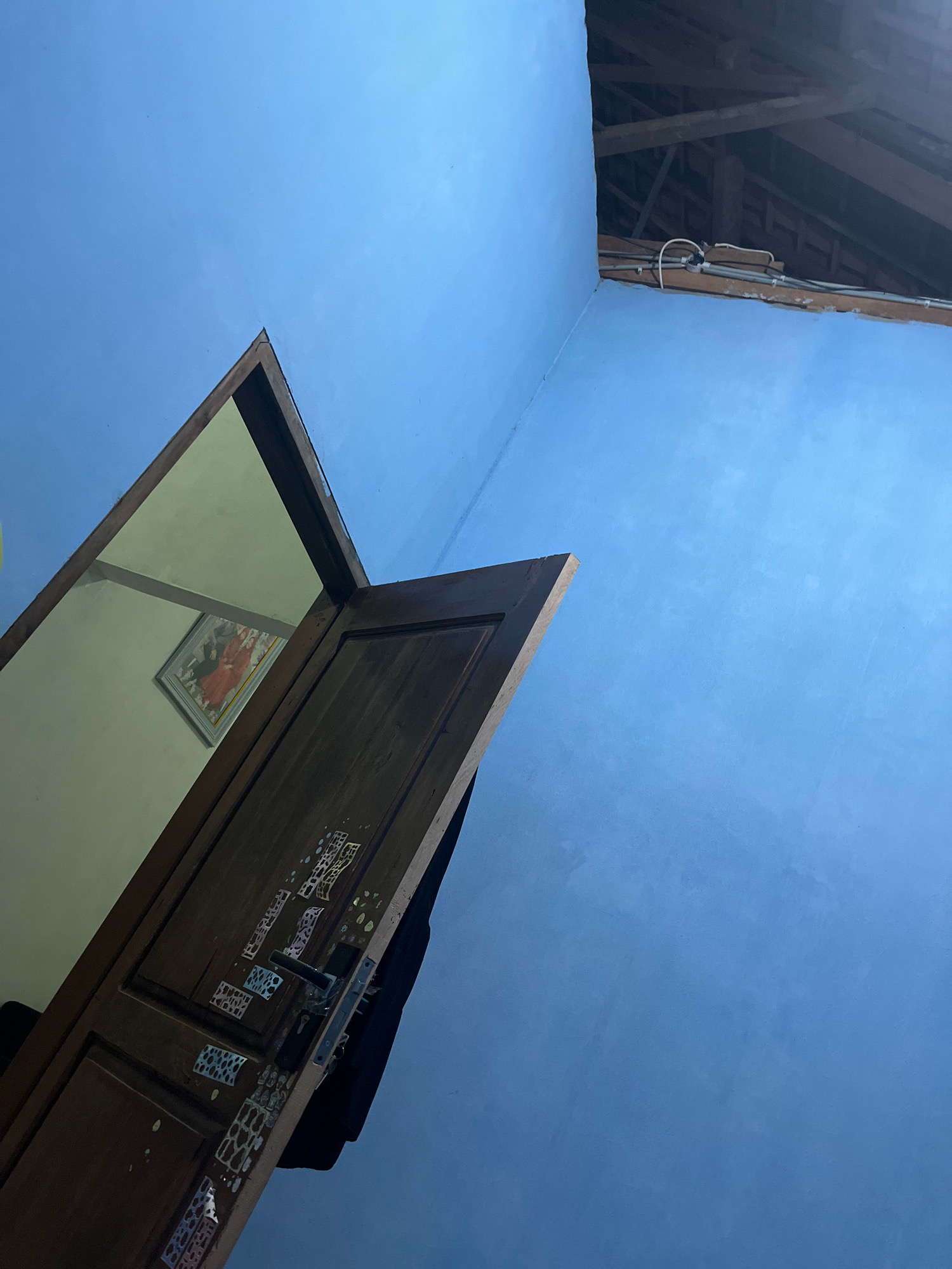 An interior scene in Kebumen, Indonesia, captured from a low angle, shows a light blue wall on the right and a dark wooden door ajar on the left. The door is partially open, revealing a glimpse into an adjacent room with a light-colored wall and a framed picture hanging on it. The picture appears to be a religious or cultural depiction, with figures in red and orange attire. The door itself is adorned with decorative metal plates. Above the wall on the right, exposed wooden roof beams and electrical wiring are visible, suggesting a rustic or unfinished architectural style. The overall lighting suggests it is either early morning or late evening.
