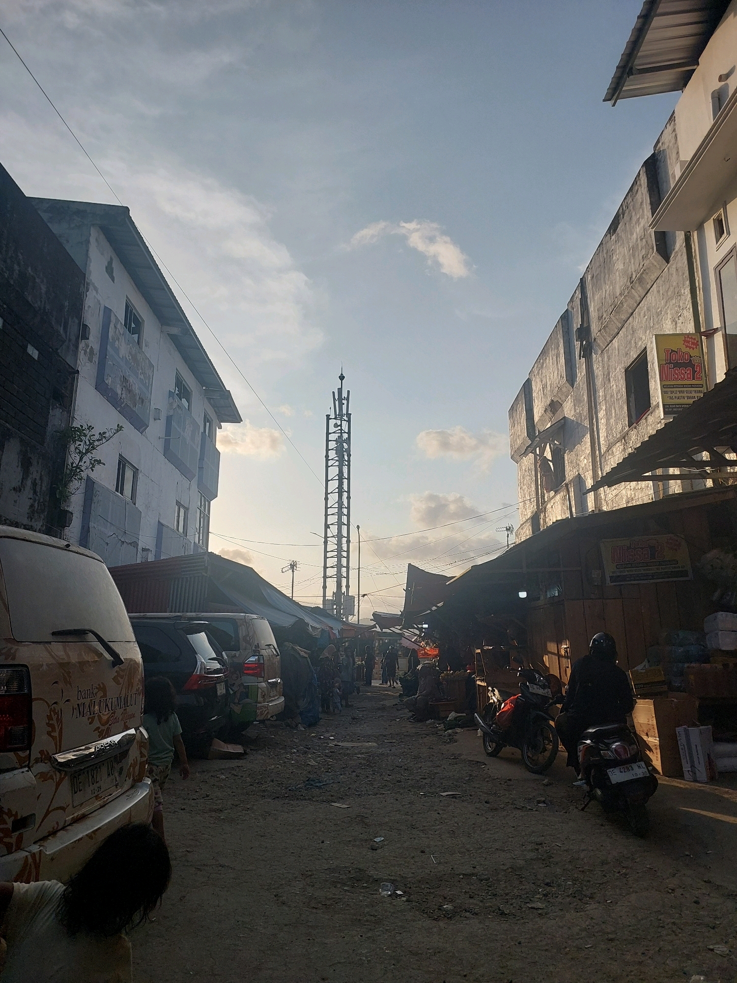 The image depicts a street scene in Ambon, Indonesia, likely a market area given the presence of stalls and vendors. On the left, a multi-story building with white and blue facades stands, partially obscured by a parked van with floral decorations and text that includes "MALUKUMALUT". Adjacent to this, a row of parked vehicles, including a dark SUV, are lined up. The street itself is unpaved and dusty, with debris scattered on the ground.
In the center of the image, a tall, lattice-like telecommunications tower rises against the sky, which is partly cloudy. To the right, another building features a grey, textured wall. Underneath a corrugated metal awning extending from this building, stalls are set up. A sign with red and yellow lettering, reading "Toko Nissa 2", is visible. Further along the right side, more stalls and piles of goods, including boxes and bags, are arranged.
Several individuals are present. In the foreground, a person is crouched down next to the decorated van. A child in a light blue shirt is walking near the parked vehicles. Further into the scene, people are seen interacting near the market stalls, some appearing to be vendors or shoppers. Two motorcycles are parked in the street; one is red and the other is dark with a rider wearing a black helmet and jacket. The lighting suggests it is either early morning or late afternoon, with long shadows cast across the ground. The overall impression is of a busy, somewhat makeshift commercial area.