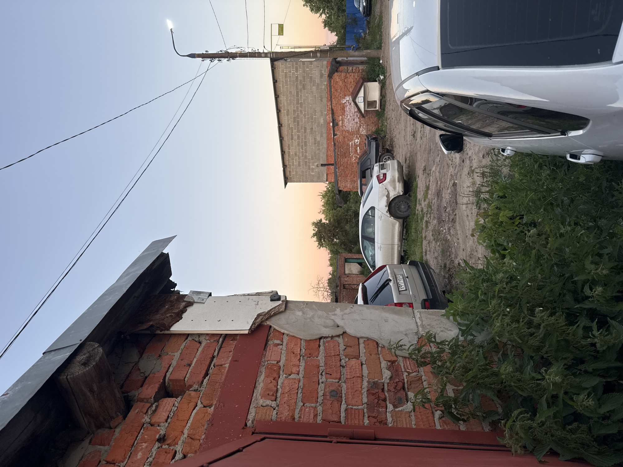 The image captures an outdoor scene, likely in Боголюбово, Russia, during what appears to be late afternoon or early evening, given the warm, soft light of the setting sun. The primary subjects are several cars parked on a dirt or gravel lot. One prominent silver car is in the foreground, its side and rear visible. Another white car is parked behind it. In the background, a brick building with a lighter colored block-built section is visible, with some signs of disrepair, particularly around the roofline. Trees and greenery are present, suggesting a somewhat rural or suburban environment. Overhead, a street lamp is illuminated, and electrical wires stretch across the sky. A portion of a red-tiled roof or wall is in the lower part of the frame, adding to the impression of a residential or industrial area. There is no clear activity or event taking place; the cars are parked, and the scene appears static. Notable details include the construction materials of the buildings and the condition of the foreground roof, which shows exposed wood and crumbling plaster. The sky is clear, indicating fair weather. A yellow sign with text is visible on the building in the background, but the text is not legible. A license plate on the silver car shows the text "X977 X069".