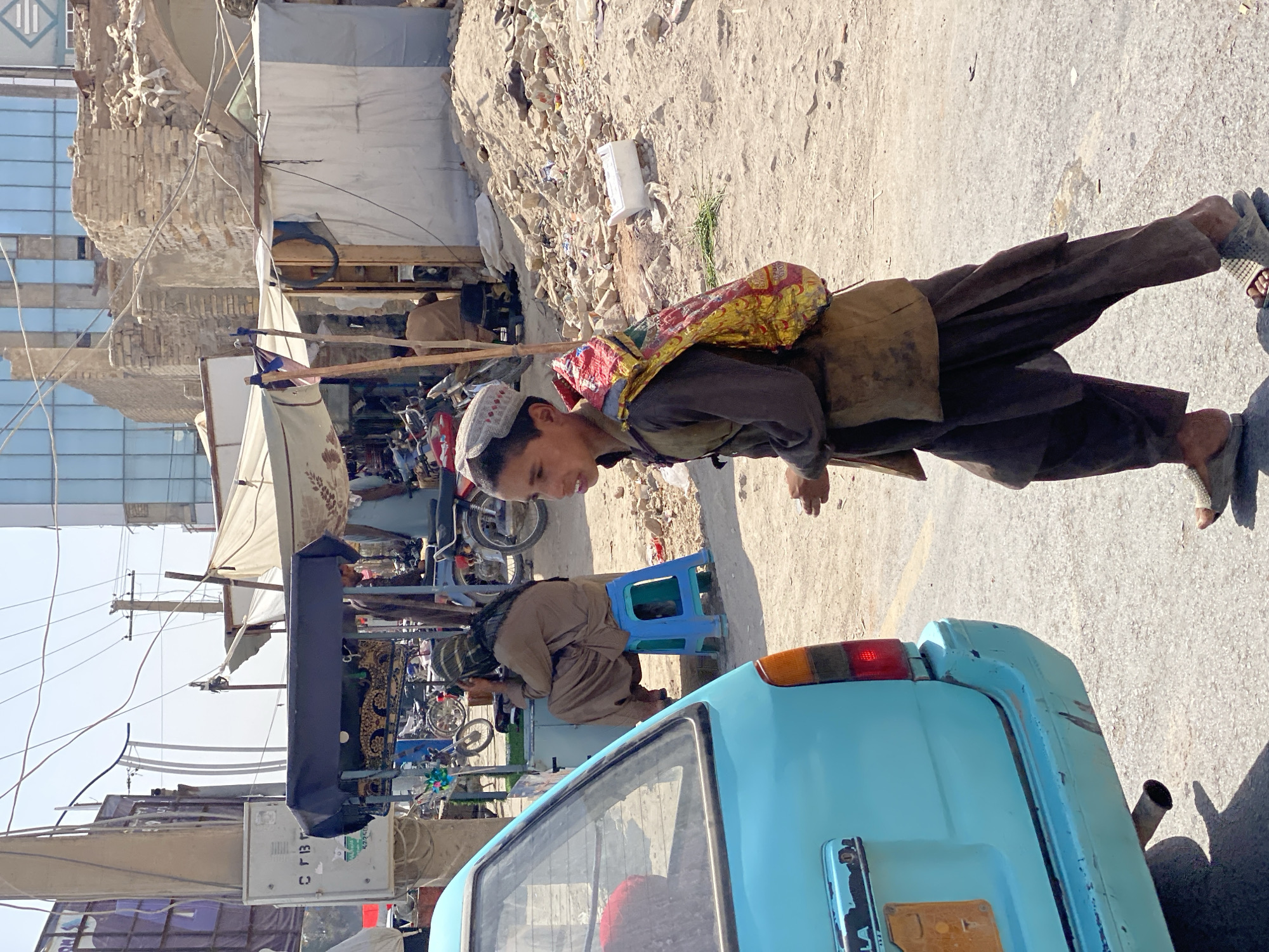 A young boy wearing a traditional cap and a vest over a brown shirt and loose pants walks on a street in Arghandab, Afghanistan. He carries a colorful bag slung over his shoulder. In the background, a light blue car is parked, and a man sits on a blue stool near a market stall. The environment appears dusty and somewhat derelict, with debris visible around the market area. The sky is bright and clear.