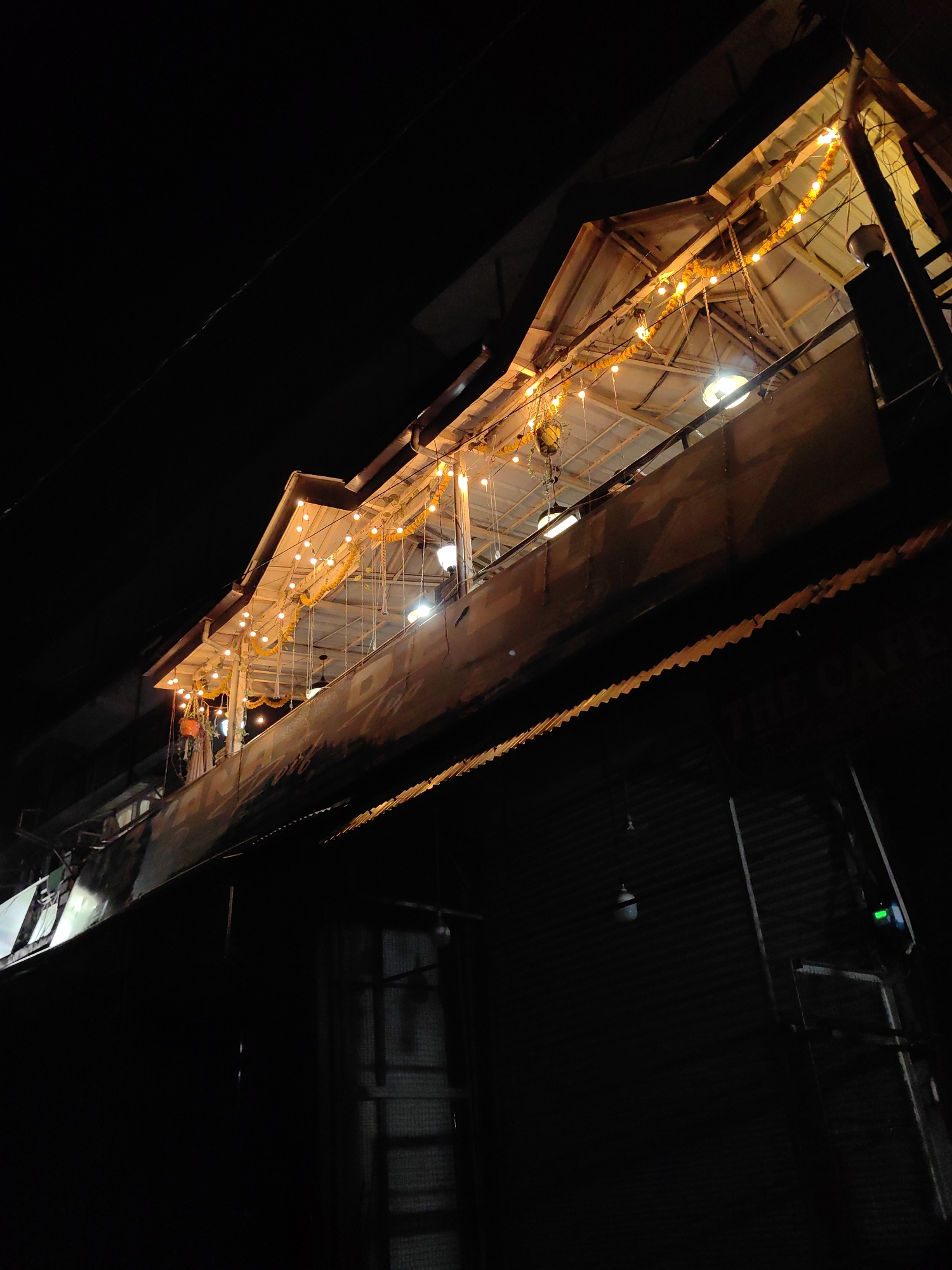 The image shows an exterior view of a building at night in Vashisht, India. The building has multiple levels with corrugated metal roofing and siding. The upper level features a balcony or walkway adorned with strings of yellow, bulb-shaped lights and floral garlands. Several light fixtures are visible under the roof of this upper level. A dark, possibly metal, structure, possibly a sign or part of the building's facade, is visible on the second story from the bottom, with some faded markings. A green indicator light is visible on the right side of the building's lower section. The overall impression is of a rustic or traditional establishment illuminated for nighttime activity.