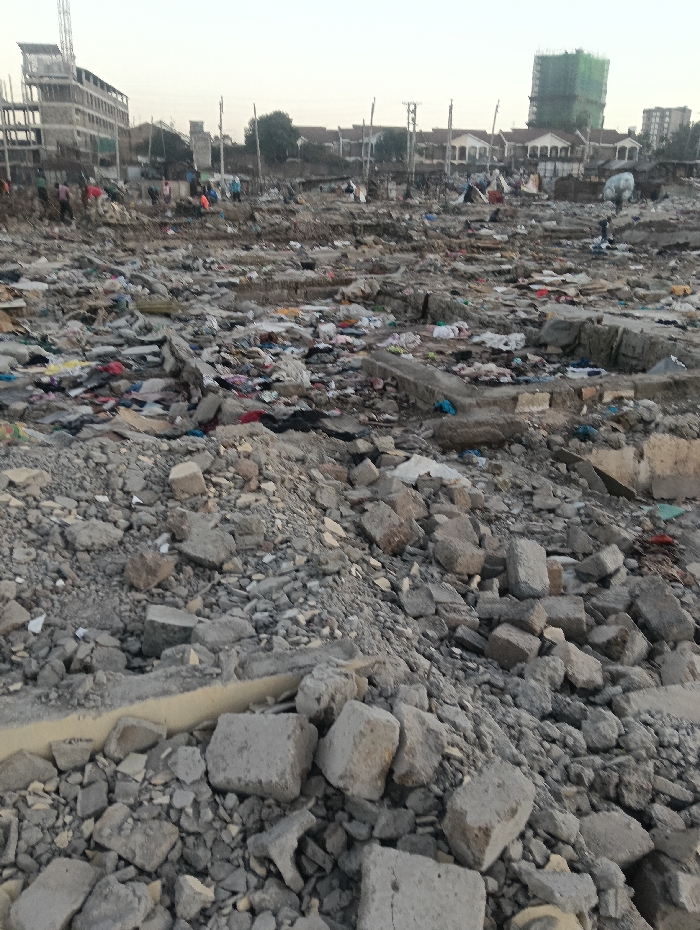 The scene depicts a desolate landscape littered with rubble, concrete fragments, and debris. This suggests a site of destruction, possibly from a natural disaster or demolition. In the background, buildings and structures stand amidst the ruin. People can be seen in the distance, navigating through the wasteland, perhaps searching or salvaging. The overall atmosphere is one of devastation and disruption.