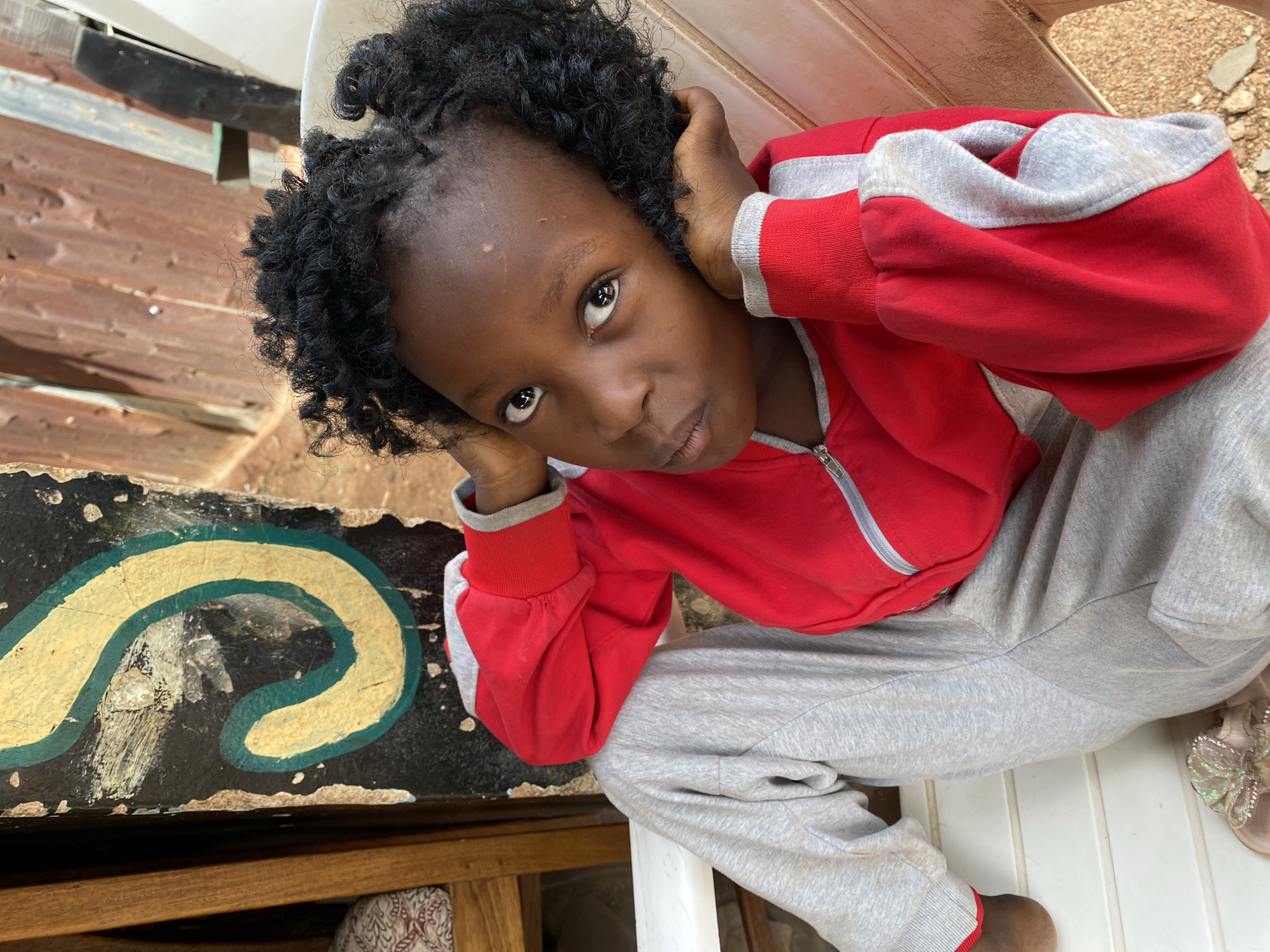 A young girl with curly black hair is captured in a medium, high-angle shot, seemingly in Kuru Karama, Nigeria. She is seated, wearing a red and grey zip-up jacket, and grey sweatpants. Her hands are near her head, with an expression looking directly at the camera. To her left is an old, dark-colored piece of furniture with a yellow and green design. The background appears to be outdoors. The setting appears casual and intimate, possibly residential.