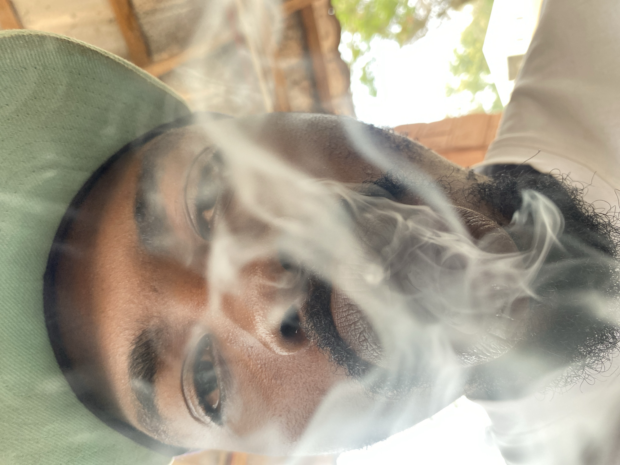 The image features a close-up, angled portrait of an individual, likely male, with dark skin and a beard and mustache. He is wearing a light-colored, possibly sage green, brimmed hat and a light-colored shirt. A significant amount of dense, white smoke billows around his face, partially obscuring his mouth, nose, and parts of his eyes, suggesting he is exhaling smoke from smoking or vaping. His eyes are open, and his expression appears neutral or relaxed. The photograph is taken from a low angle, looking upwards. The blurred background reveals bright daylight, indicating a daytime setting. Visible elements in the background include green foliage, possibly from trees, and some light-brown, textured structural elements, suggesting an outdoor or semi-outdoor location in Jos, Nigeria. No text is visible in the image.
